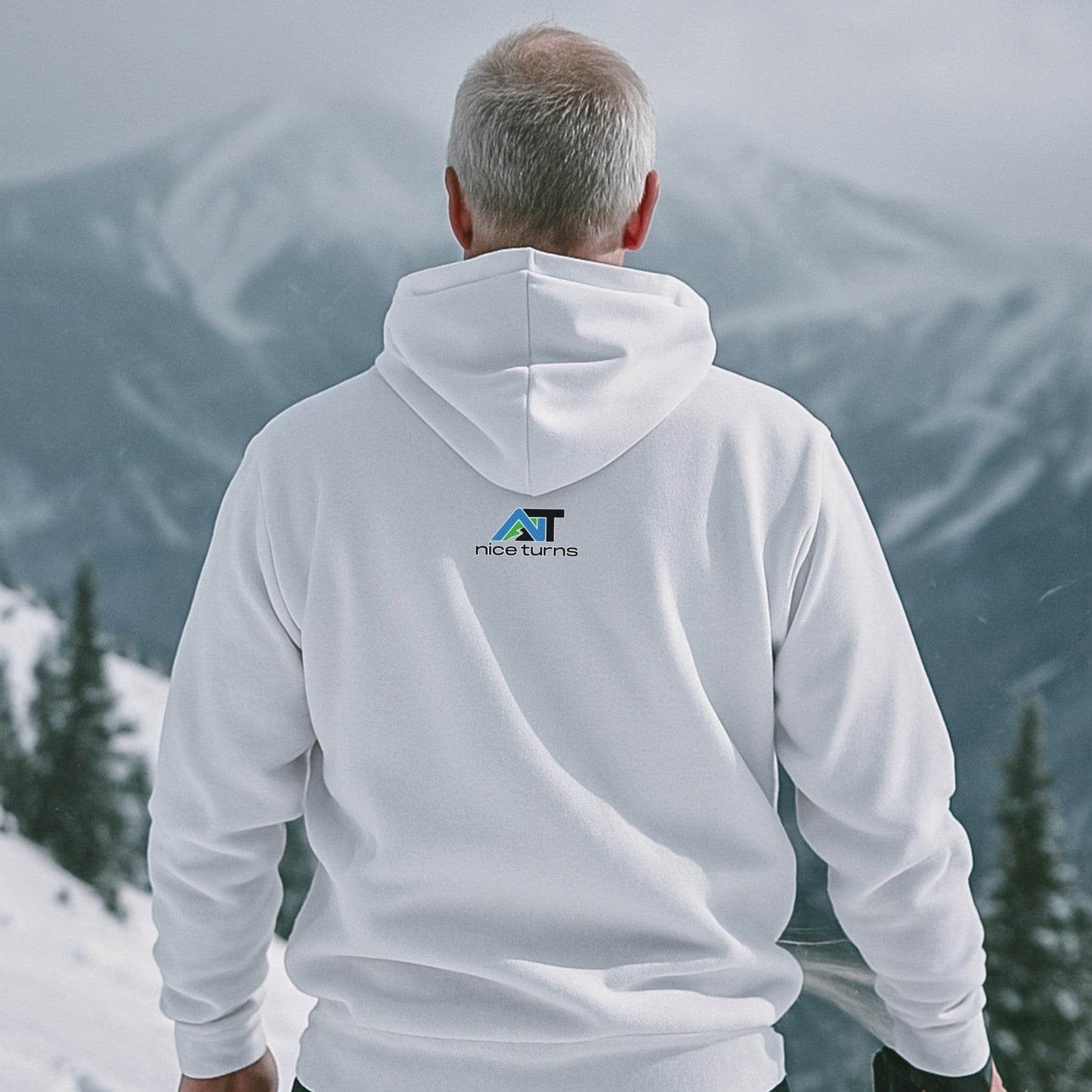 White hoodie with small nice turns logo on the back, worn by a skier looking out over a snowy alpine ridge on a cloudy day.
