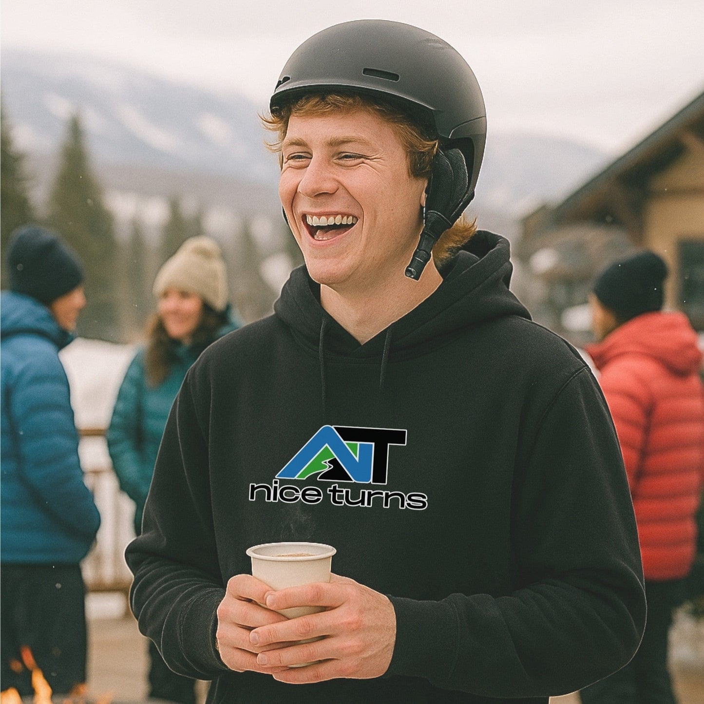 Black nice turns hoodie with call sign logo, worn by a skier in a helmet laughing by the firepit outside the lodge on a cloudy mountain day.