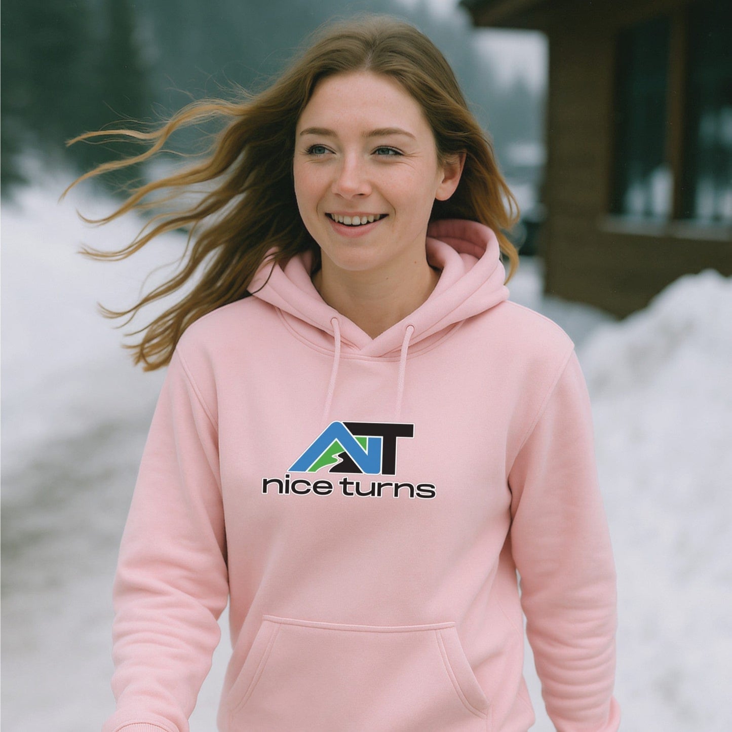 Pink nice turns hoodie with the bold call sign logo, worn by a smiling skier walking past a snowy cabin at the base of the mountain.