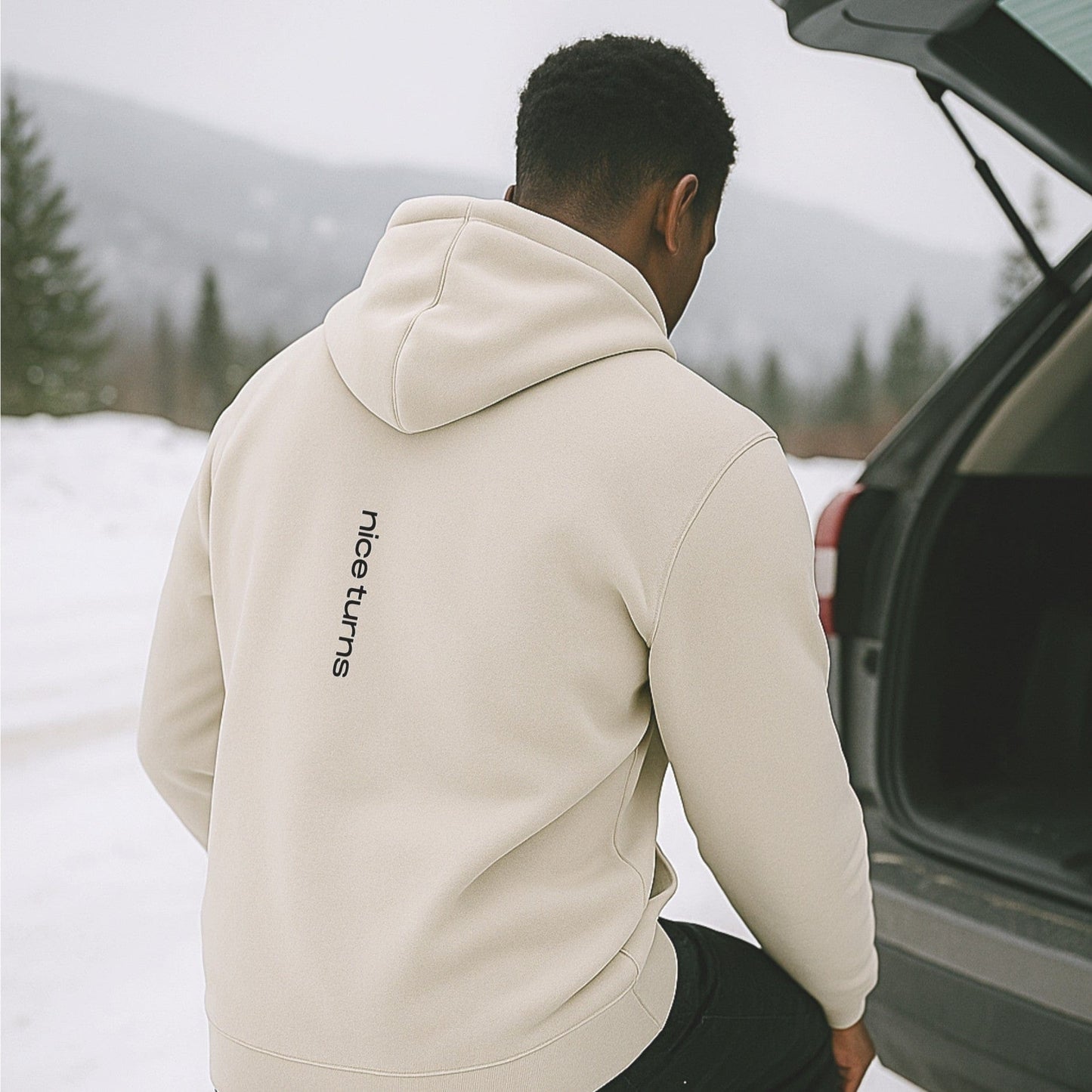 Sand-colored nice turns hoodie with vertical back print, worn by a skier gearing up at the tailgate in a snowy mountain parking lot.