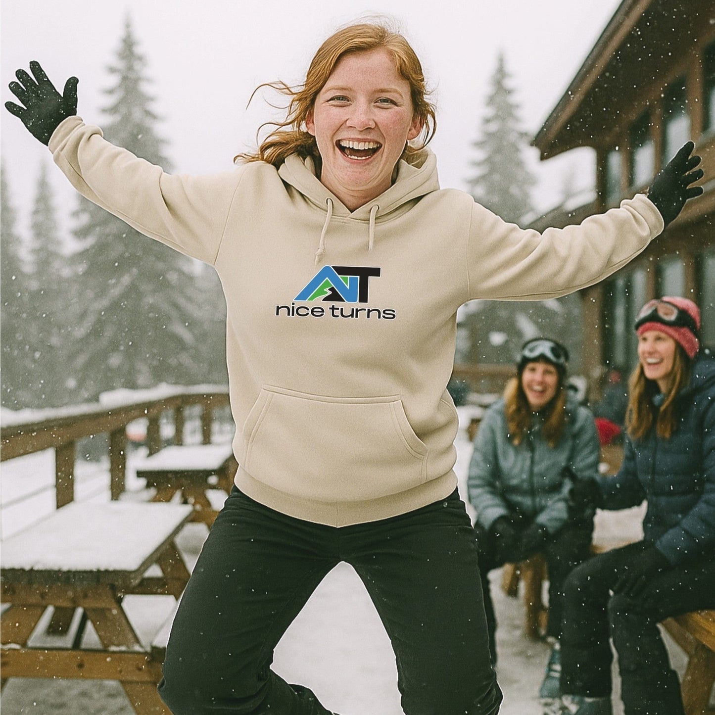 Sand nice turns hoodie with mountain logo, worn by skier mid-jump outside a snowy lodge with friends laughing on a ski deck.