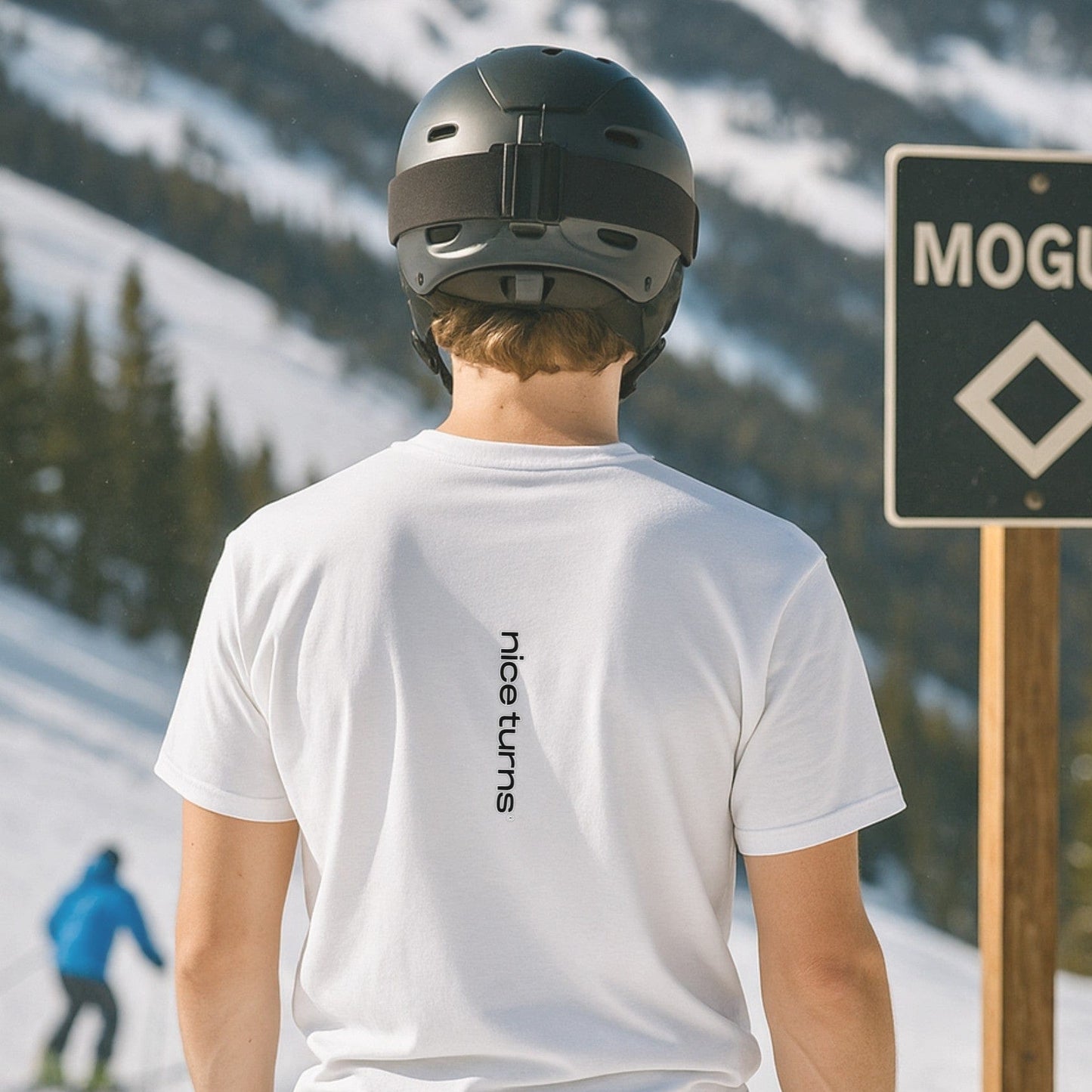 Skier wearing white T-shirt with vertical “nice turns” logo on back, standing near mogul trail sign on snowy mountain slope.