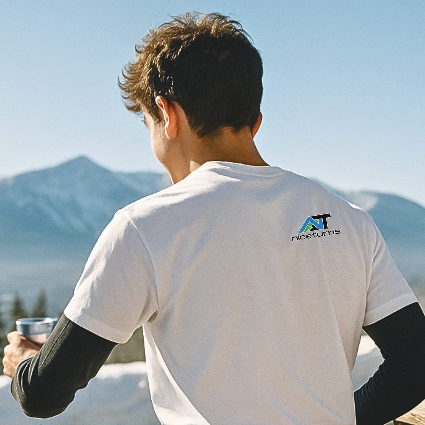 Skier holding a cup at mountaintop overlook in white No Lift Left Behind tee, logo printed near shoulder blades, morning sun and snow-covered peaks behind.