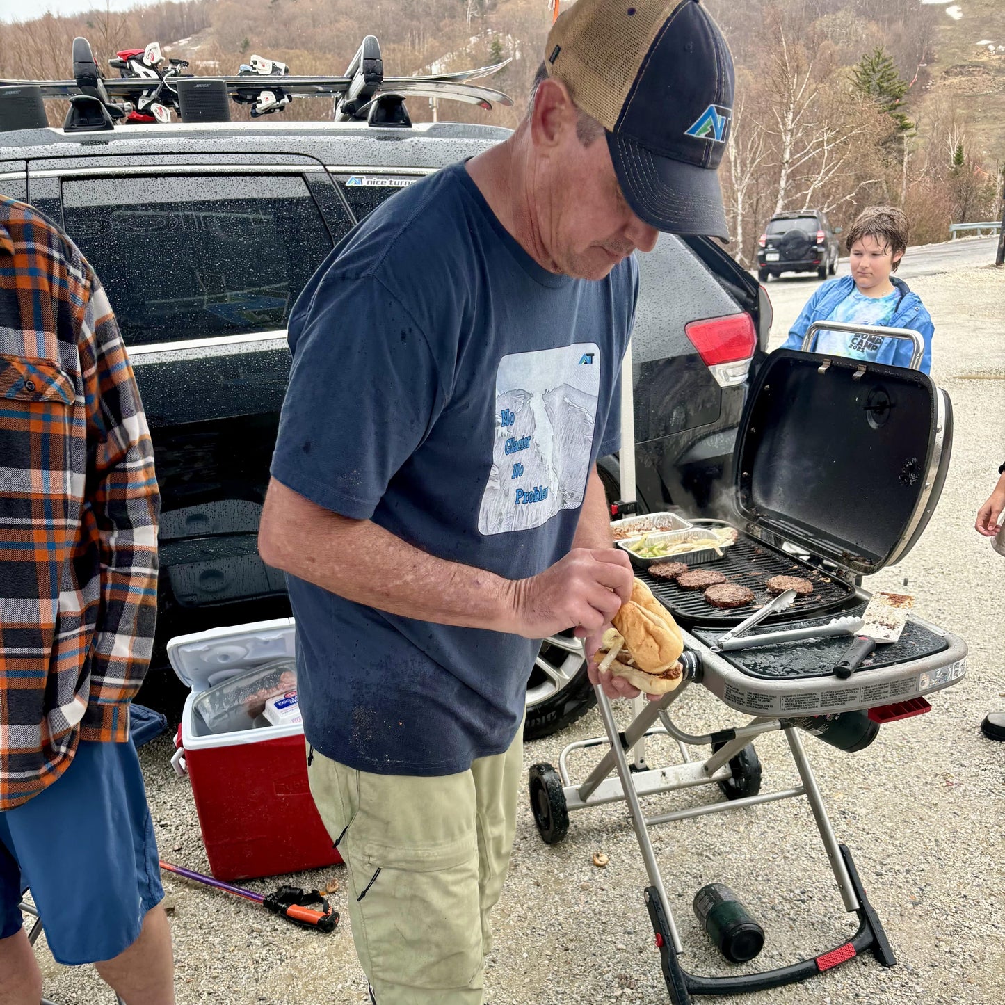 skier in navy blue no glacier no problem T-shirt grilling burgers at spring tailgate in ski lot