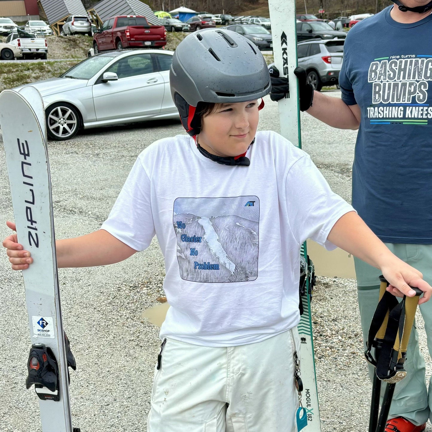 young skier in white no glacier no problem T-shirt holding skis in muddy spring parking lot