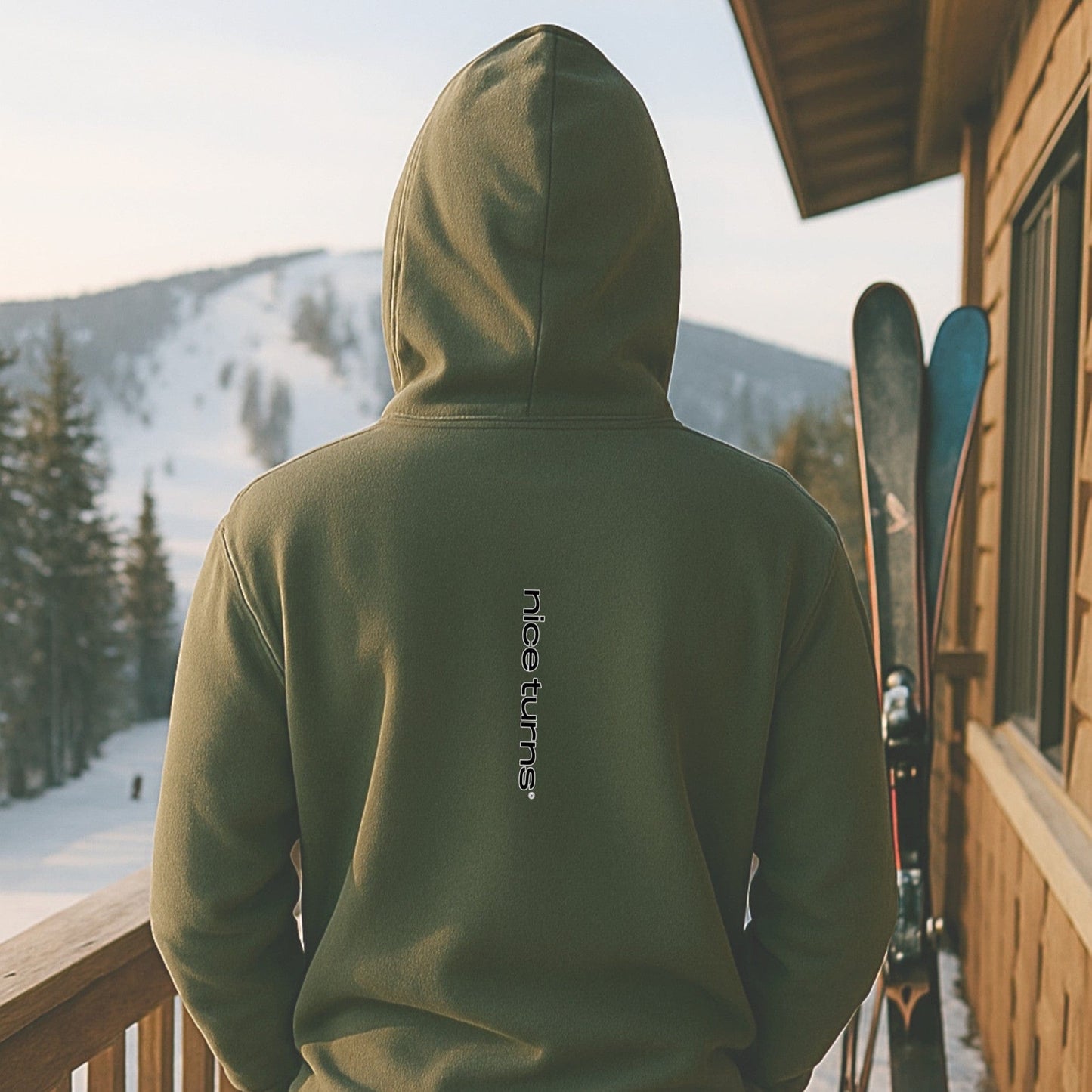 Military green hoodie with vertical nice turns logo on the back, captured on a ski lodge deck overlooking a quiet snowy mountain trail.