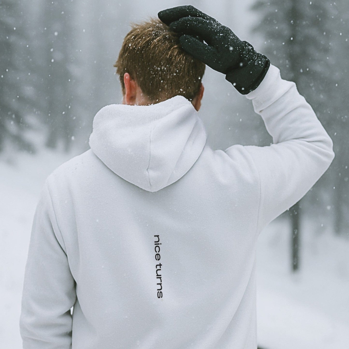 White hoodie with vertical nice turns logo on the back, shown in a snowy forest with falling flakes and soft winter light.