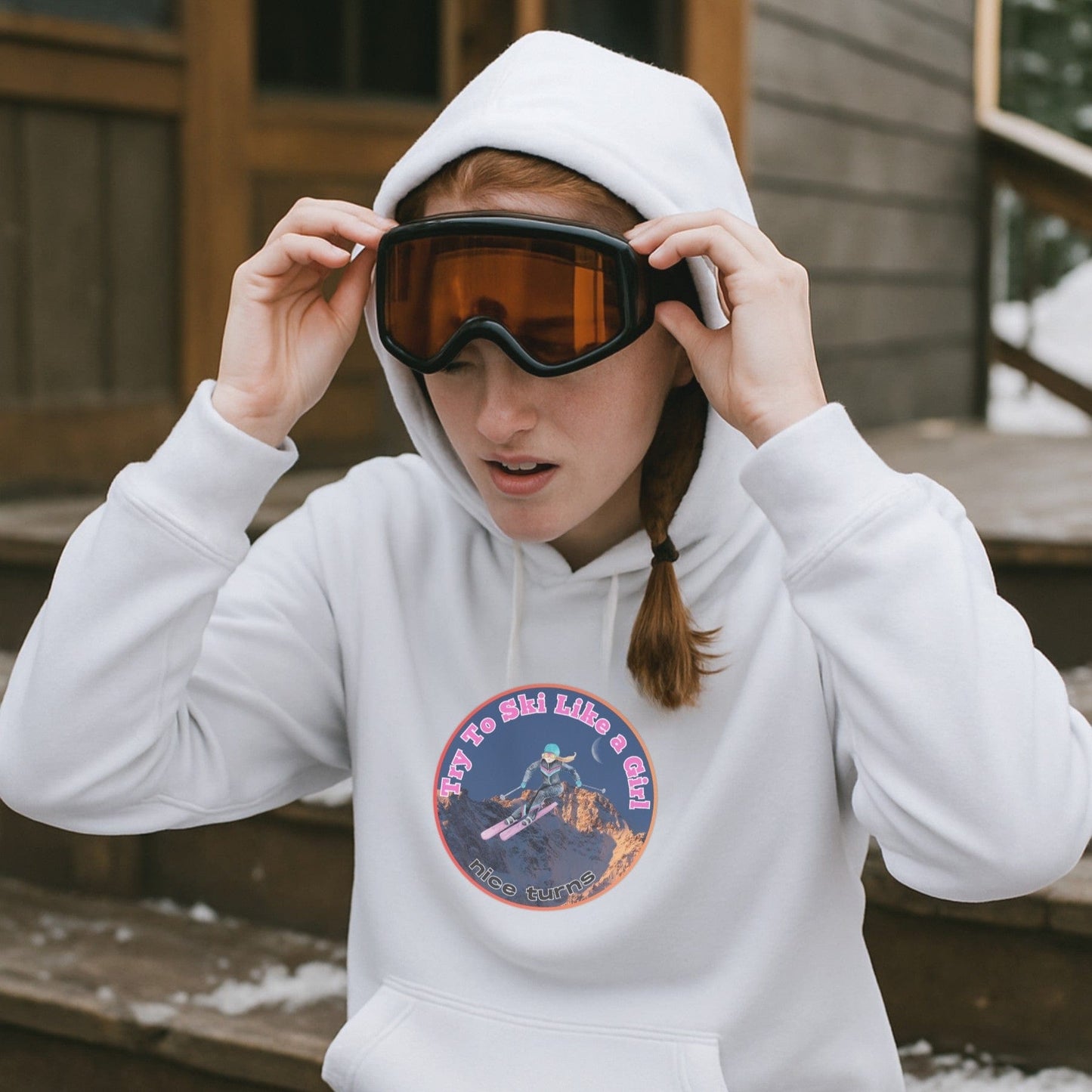 White hoodie with Try to Ski Like a Girl design, worn by skier adjusting goggles on a wooden porch after a run in the snowy woods.