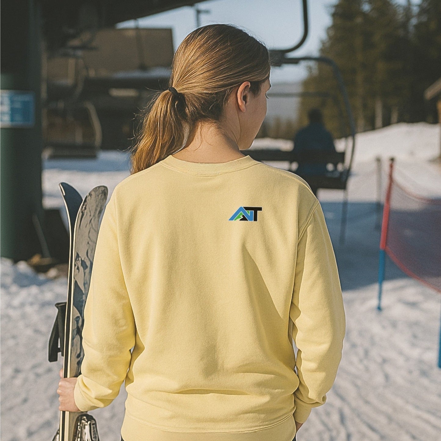 Back view of a yellow crewneck sweatshirt with AT logo, worn by skier holding poles near a lift at a snowy mountain base.
