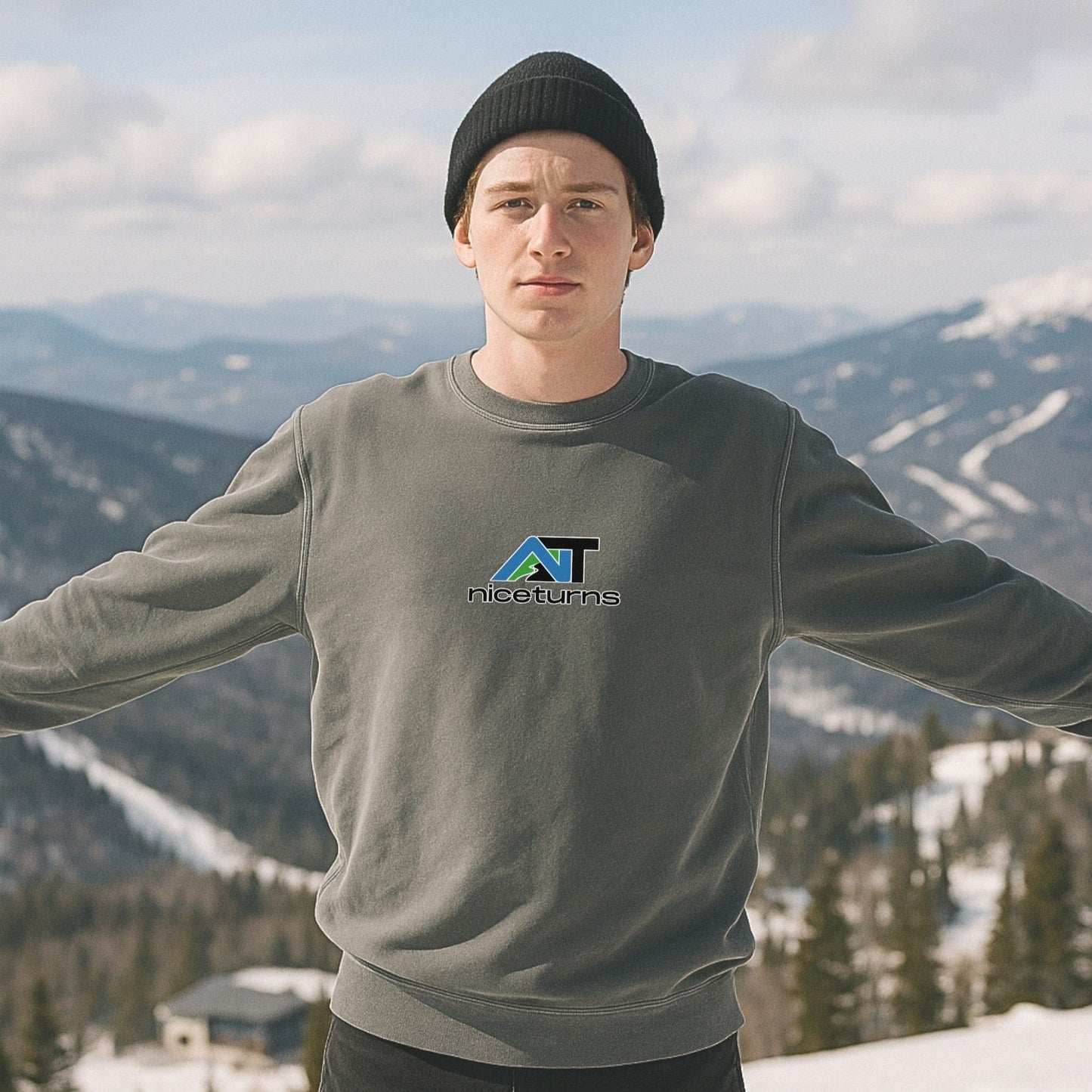 Grey crewneck sweatshirt featuring the nice turns AT logo on the chest, worn on a scenic mountain overlook with snowy slopes behind.