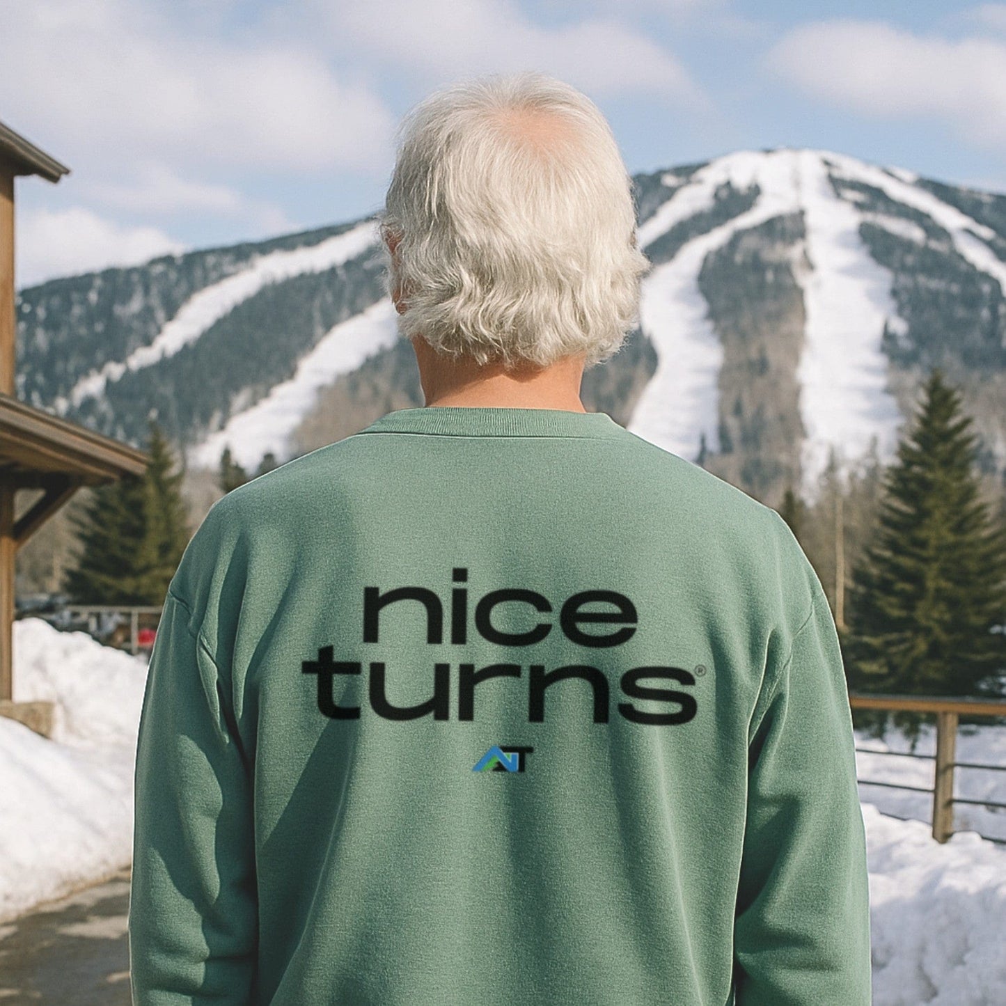 Light green crewneck sweatshirt with large nice turns logo on the back, worn at the base of a ski hill with snowy trails in view.