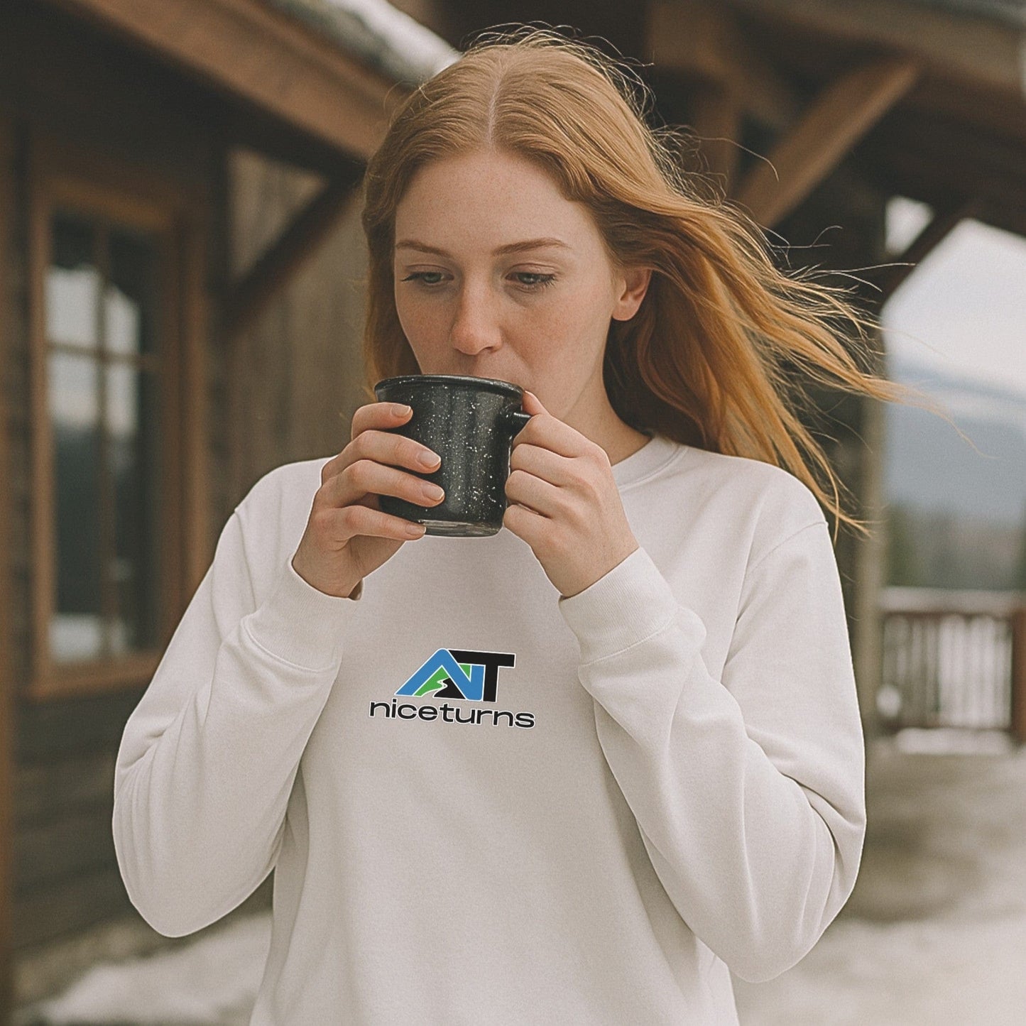 White crewneck sweatshirt with nice turns logo on the chest, worn outside a rustic ski lodge as wind blows and coffee warms the hands.