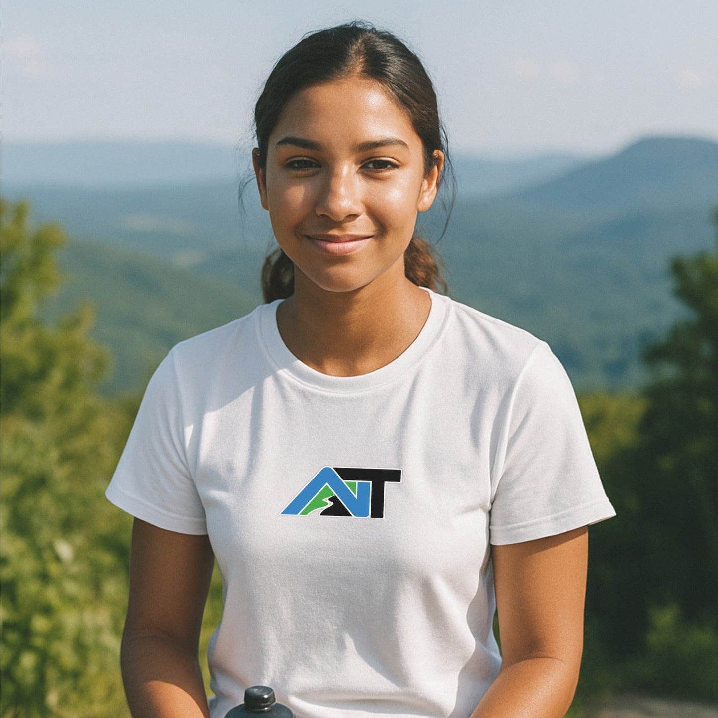 White nice turns Summit Style T-shirt with mountain logo, worn on a sunny overlook with rolling green hills in the background.