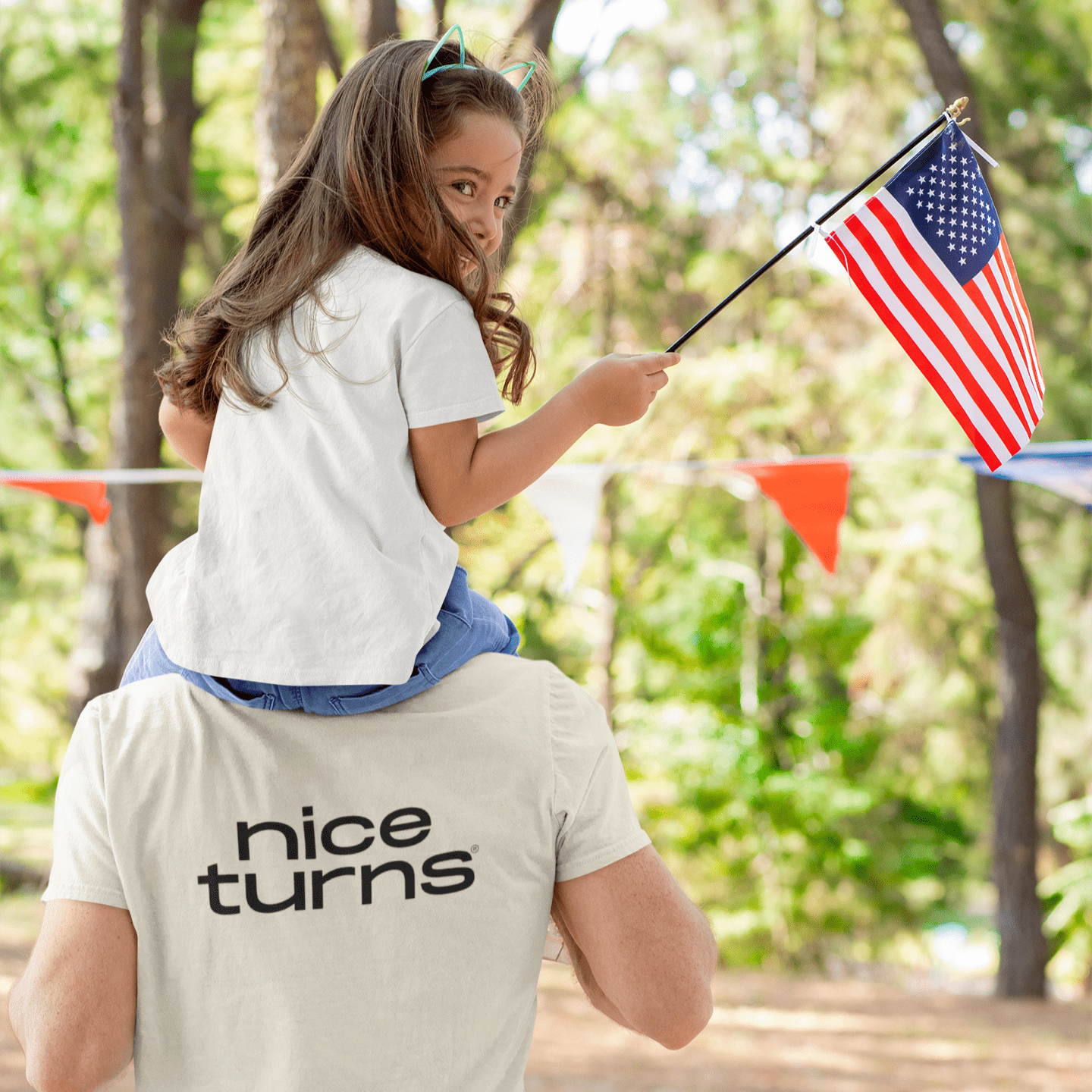 Cream nice turns T-shirt with bold back logo, worn at a summer forest gathering with a child on shoulders holding an American flag.