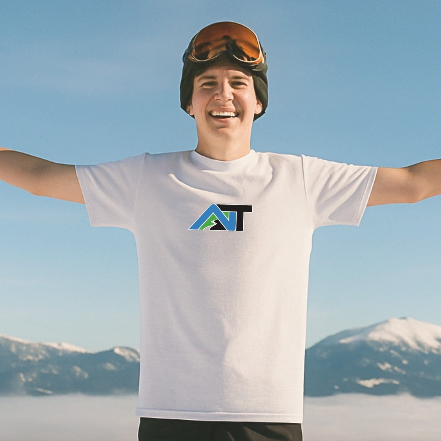 Smiling skier wearing Summit Swagger T-shirt in white stands arms outstretched on bluebird day with snow-covered mountain peaks behind.