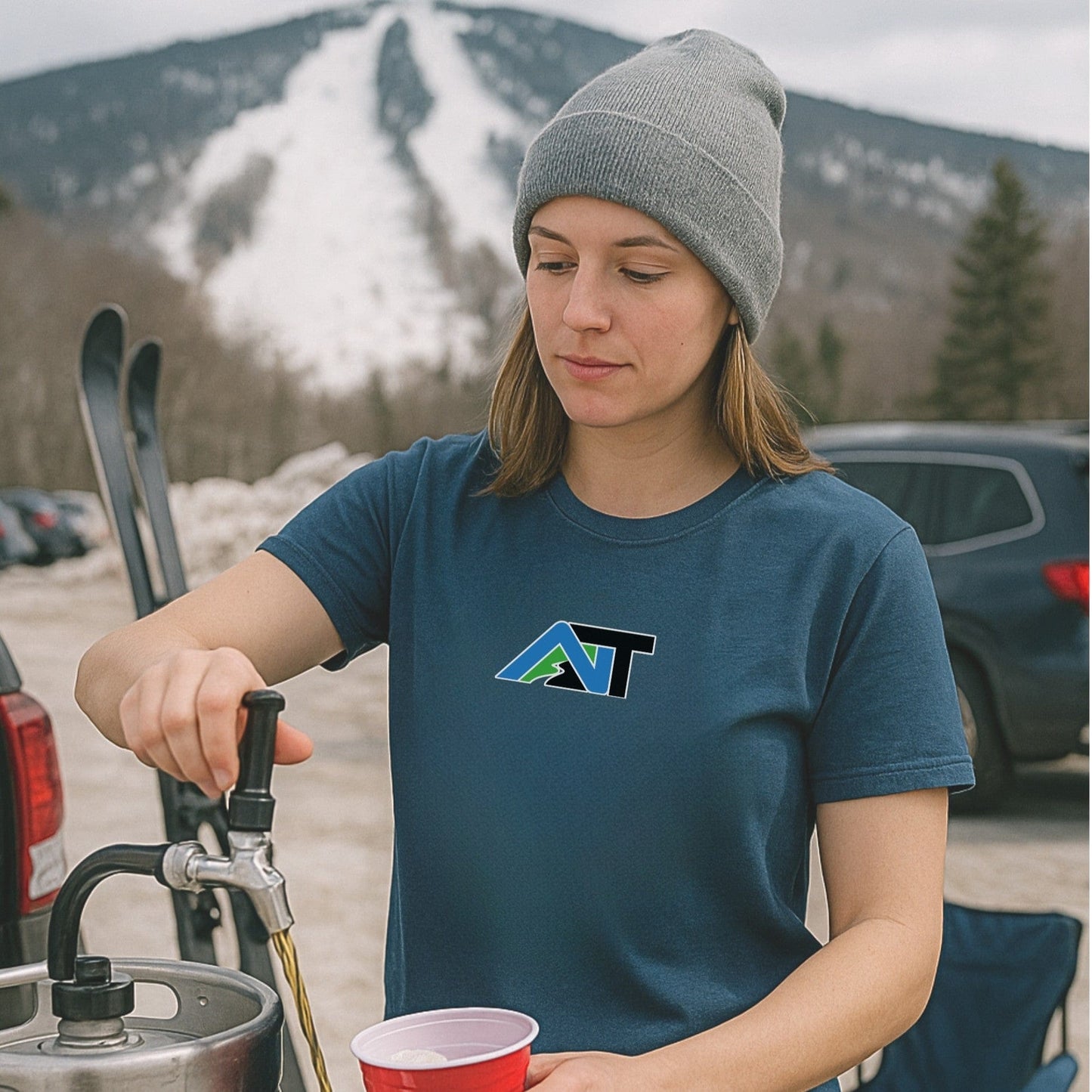 Woman wearing Summit Swagger T-shirt in navy pours beer from a keg at mountain ski lot with snowy trails and parked cars in background.