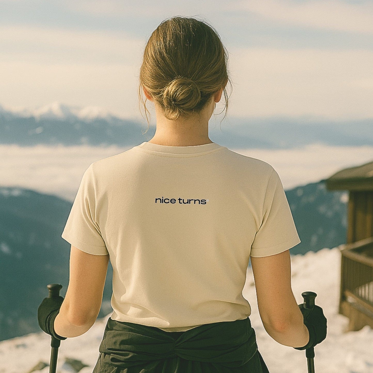 Natural color Ski Bum Skiing T-shirt with “nice turns” back print, seen from behind at a mountain overlook with poles in hand.
