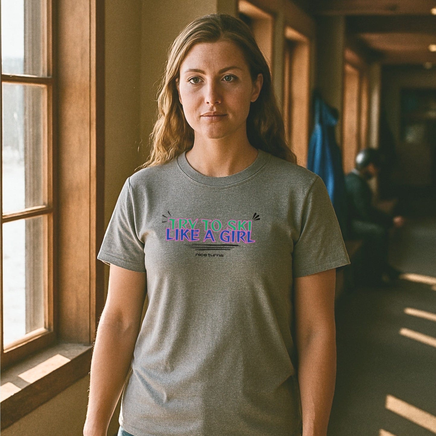 Heather gray t-shirt with the “TRY TO SKI LIKE A GIRL” slogan in neon colors, worn inside a lodge hallway with light streaming through windows.