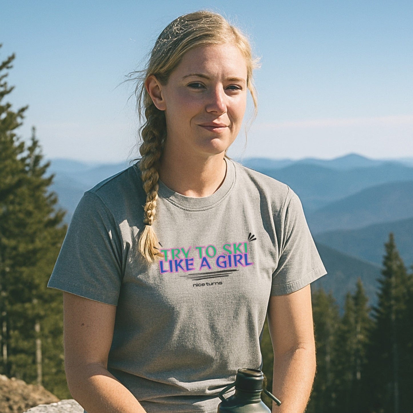 Heather gray t-shirt with “TRY TO SKI LIKE A GIRL” in bold neon text, worn at a scenic mountain overlook surrounded by evergreens and ridgelines.