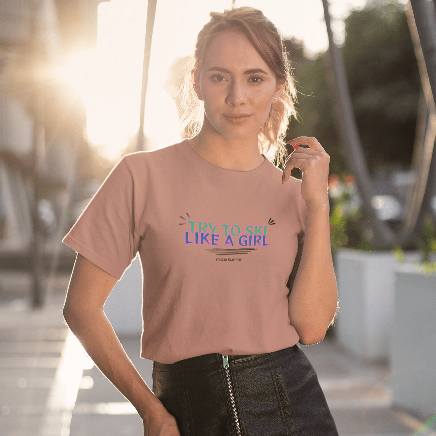 Woman wearing a pink “try to ski like a girl” nice turns tee in soft sunset light, standing casually on a city sidewalk.