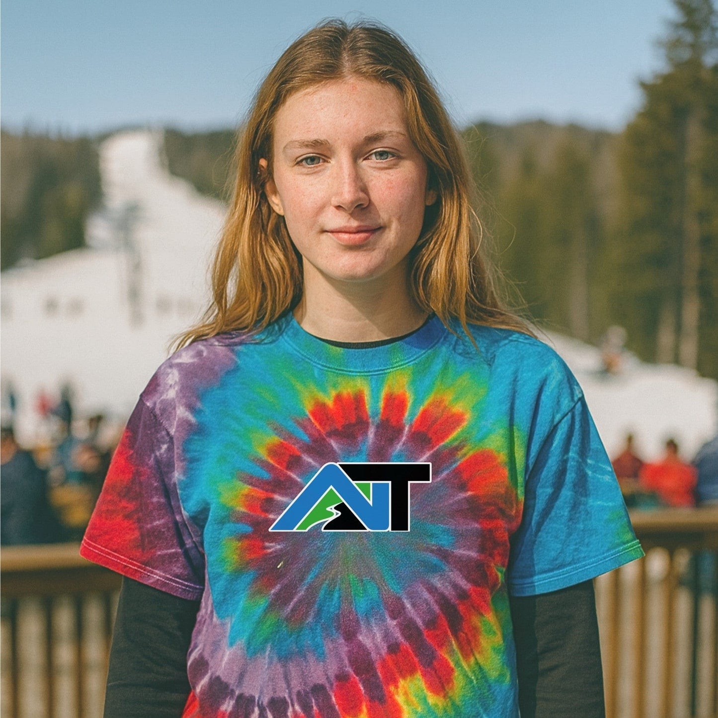 Tie-dye AT logo tee worn on a sunny spring ski day, with lifts running and crowds gathering at the base for après.