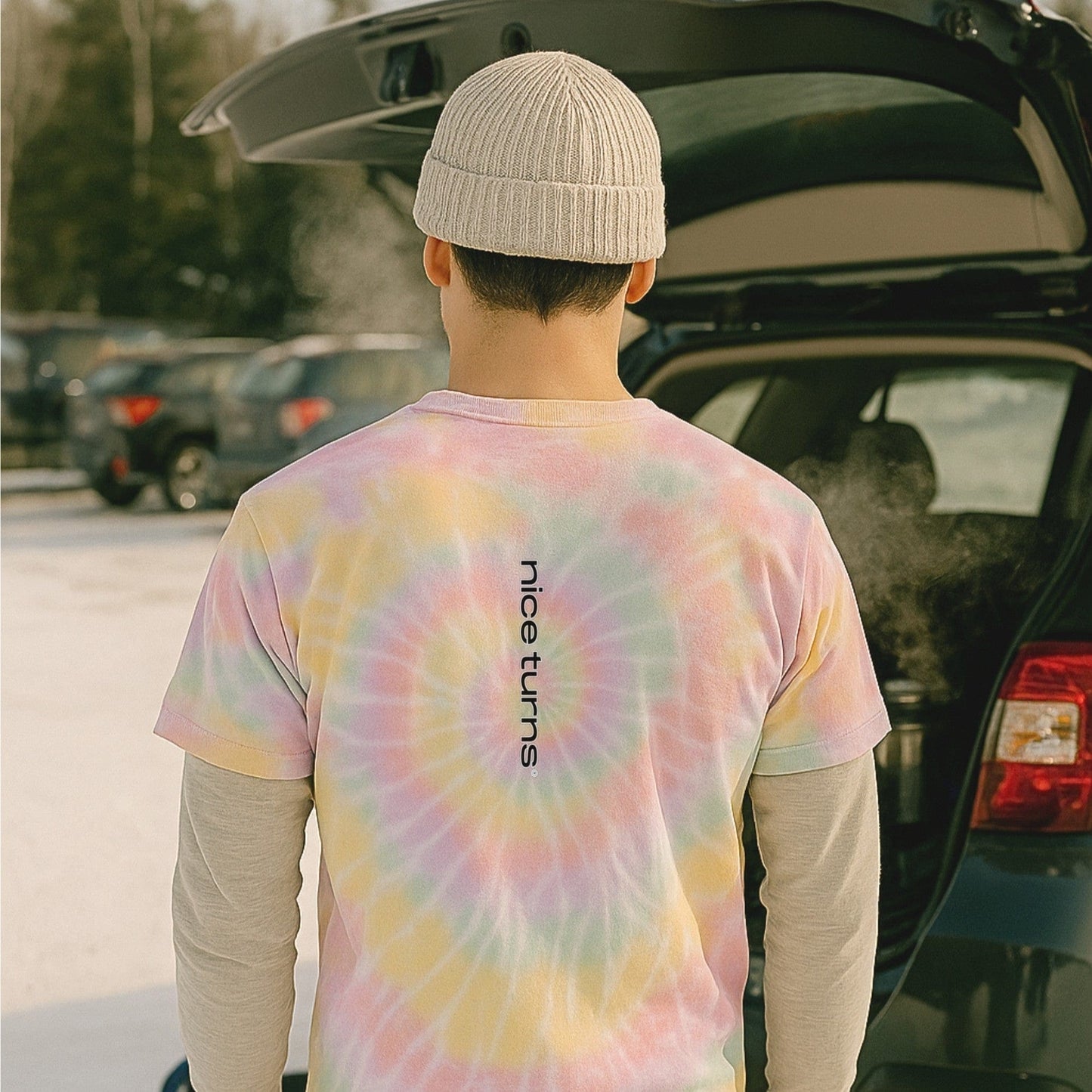 Pastel tie-dye tee with vertical nice turns logo, worn in a snowy ski parking lot with steam rising from an open car hatch.