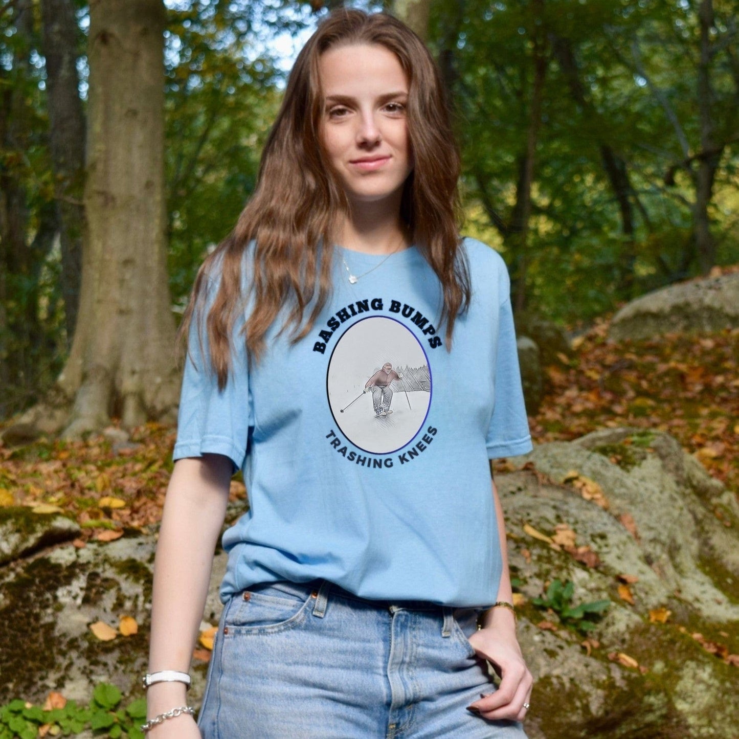 Woman wearing a light blue Bashing Bumps Trashing Knees t-shirt in a wooded early fall setting with leaves and moss-covered rocks.