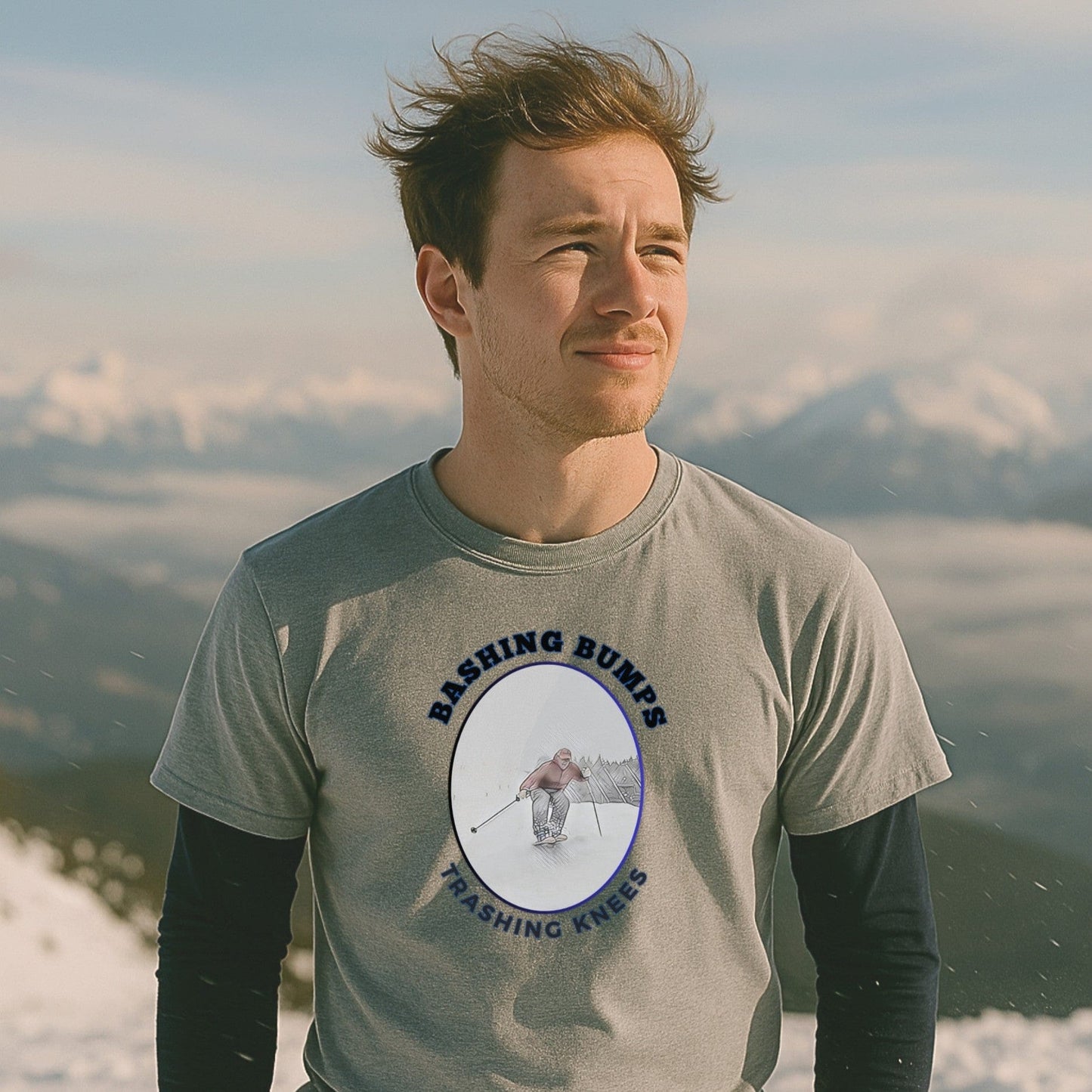 Man wearing a gray Bashing Bumps Trashing Knees t-shirt on a snowy mountain with soft light and a view of distant alpine peaks.