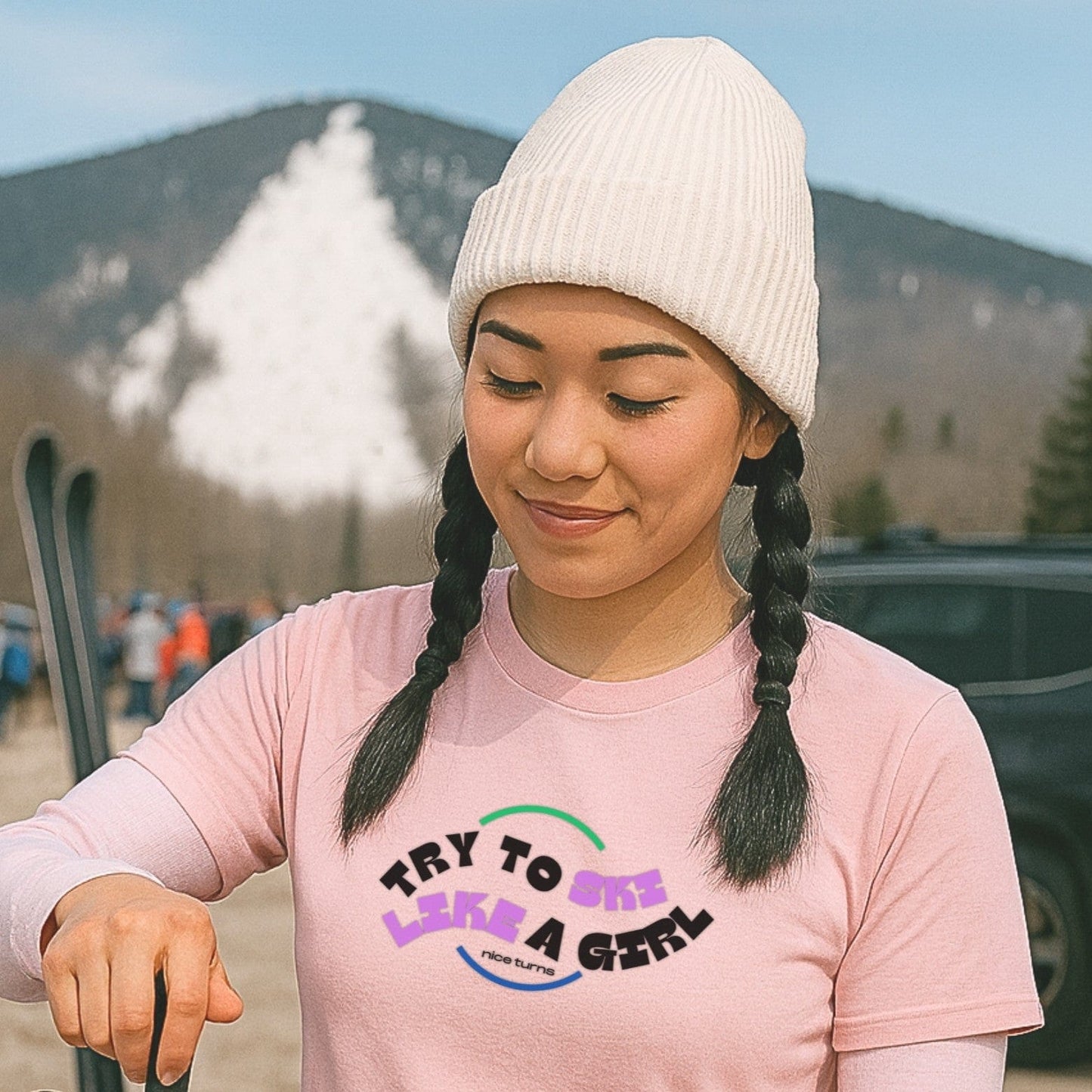 Light pink t-shirt with “Try to Ski Like a Girl” graphic from nice turns, worn in a ski parking lot with snow-covered trails in the background.