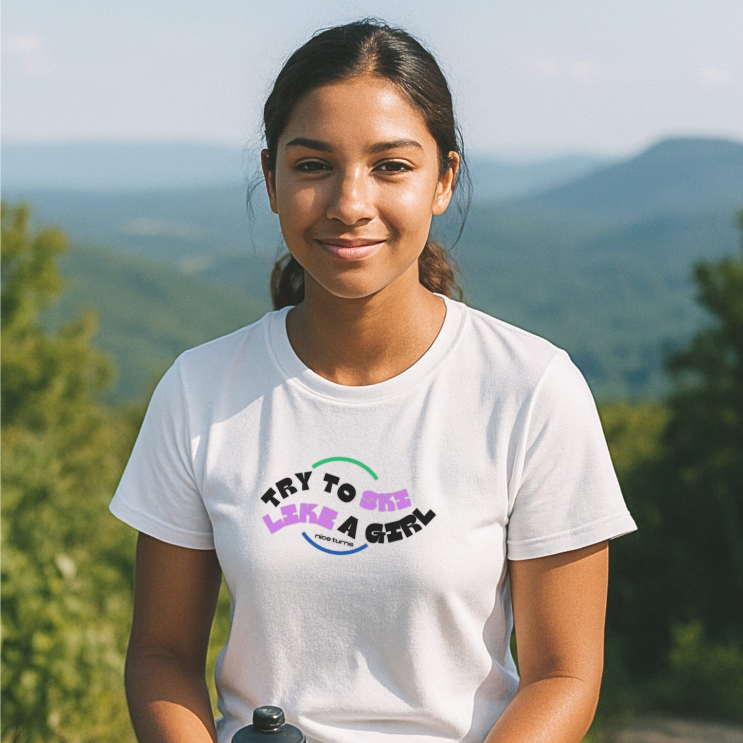 White t-shirt with “Try to Ski Like a Girl” from nice turns, worn on a sunny day overlooking a mountain valley with soft green ridgelines.