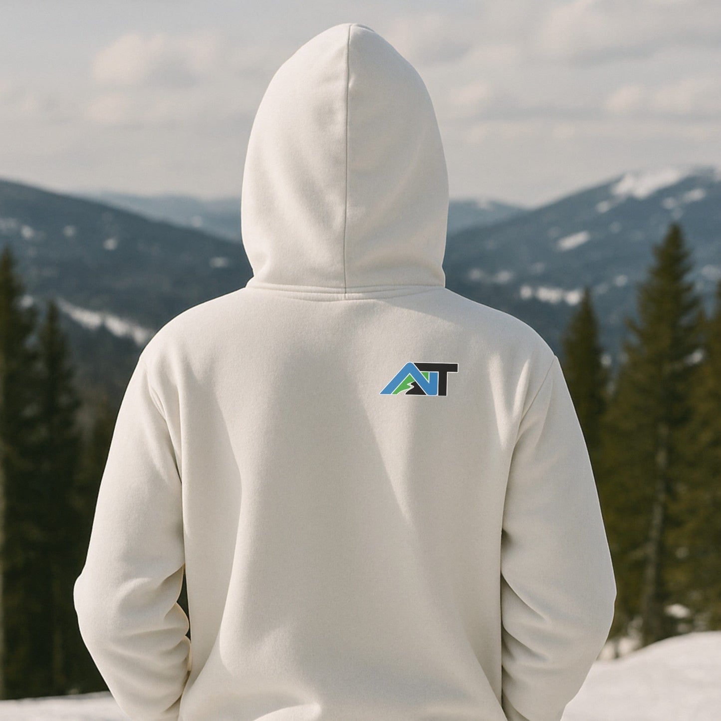 White hoodie with mountain-style AT logo on the back, worn on a snowy ridgeline with pine trees and distant alpine views.