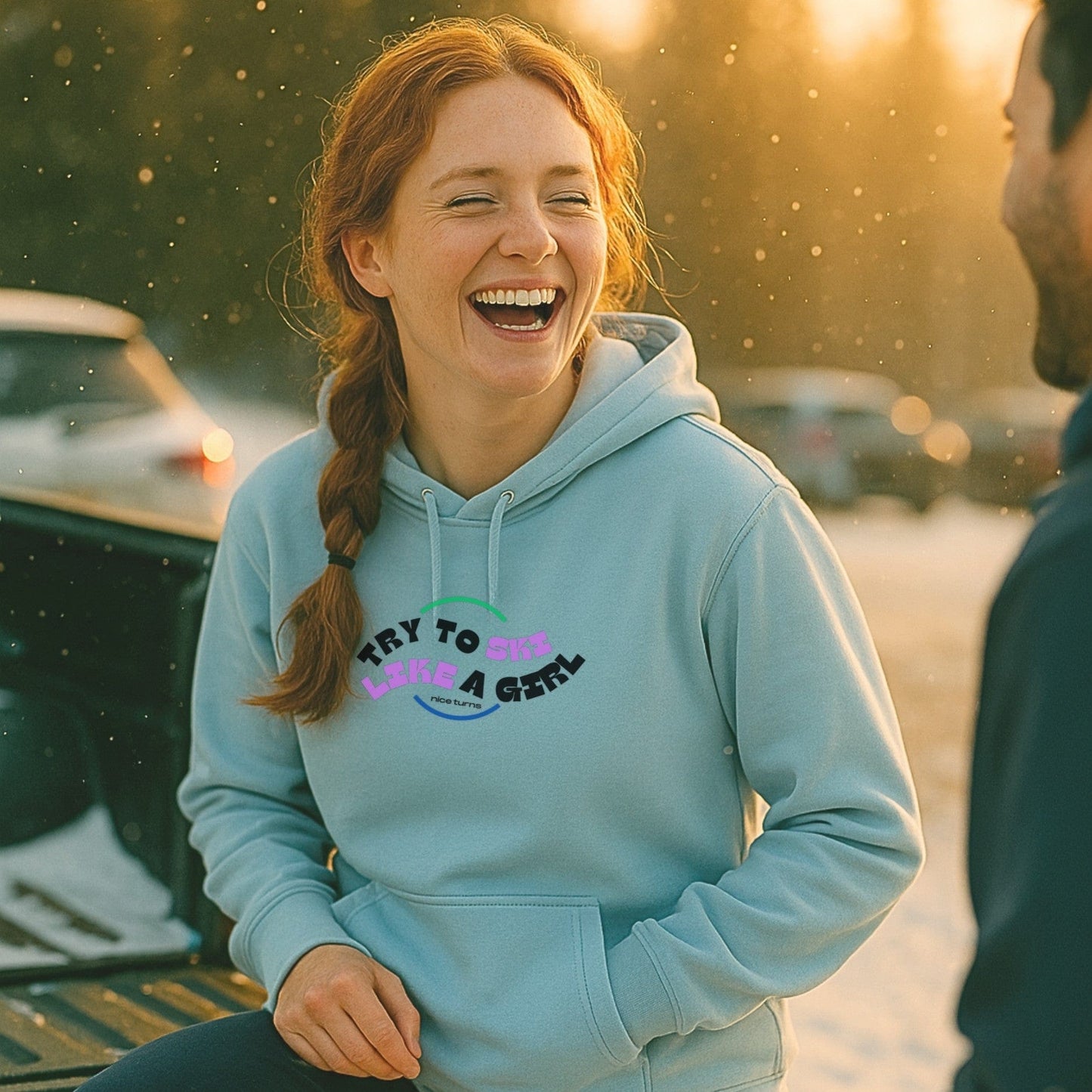 Light blue hoodie with “try to ski like a girl” arc design worn at sunset in a ski parking lot. Laughter, snowflakes, and good vibes.
