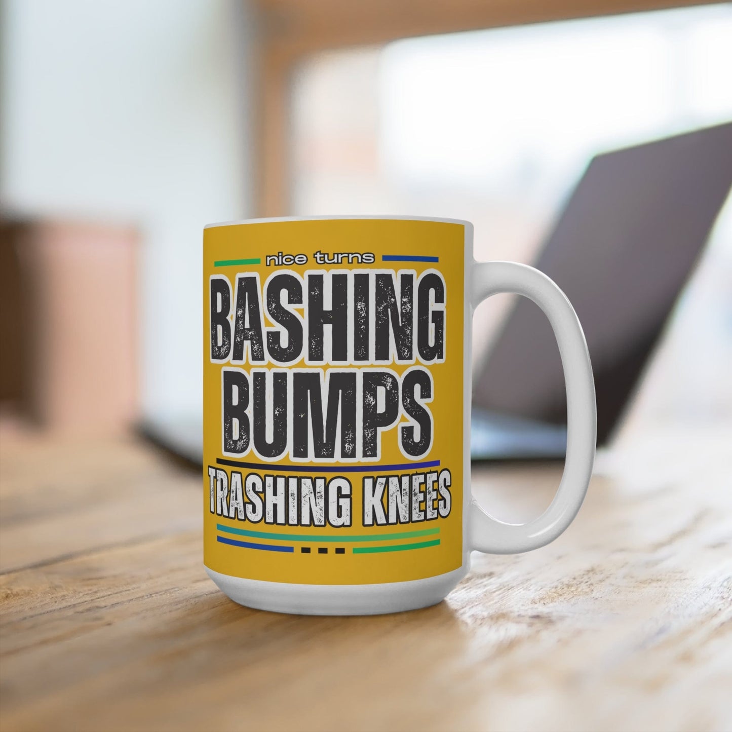 Bashing Bumps 15 oz coffee mug, yellow background, on wood desk with blurred laptop in background; bold “Trashing Knees” ski graphic in focus.