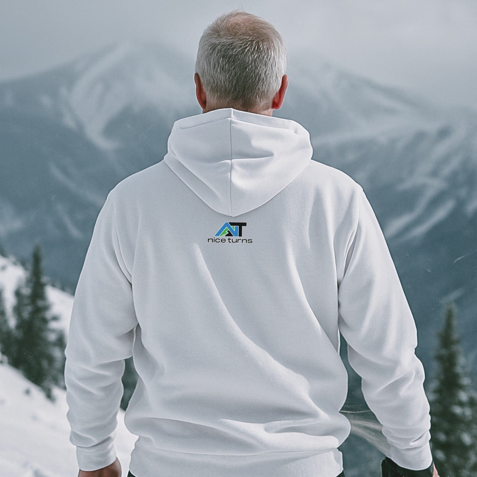 White hoodie with small nice turns logo on the back, worn by a skier looking out over a snowy alpine ridge on a cloudy day.