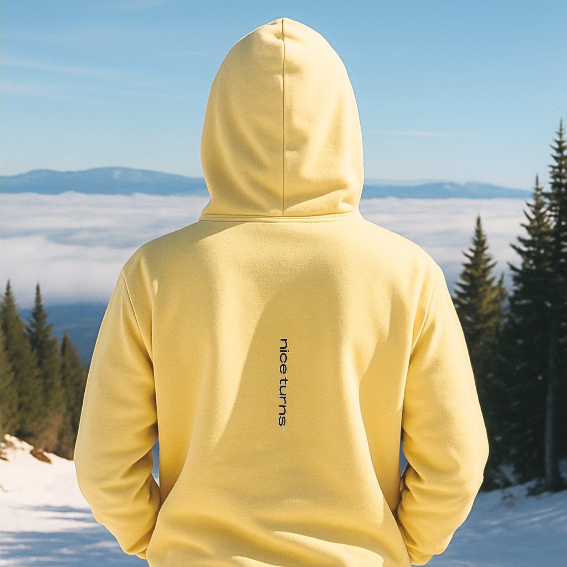 Back view of hazy yellow hoodie with vertical nice turns logo, captured on a sunny ski trail above the clouds.