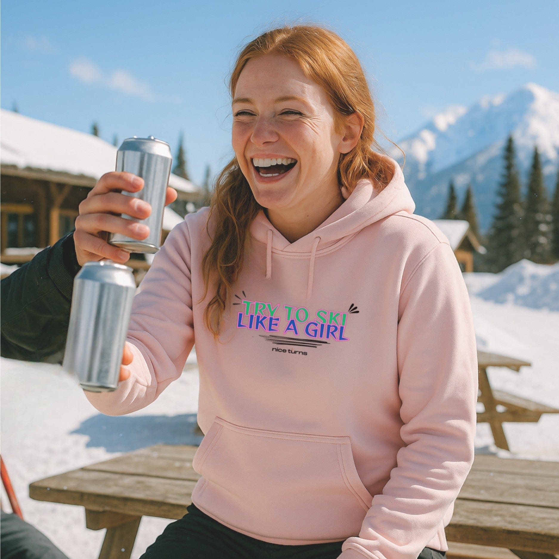 Light pink hoodie with try to ski like a girl design from nice turns, worn après-ski at a snowy lodge picnic table with mountain views.