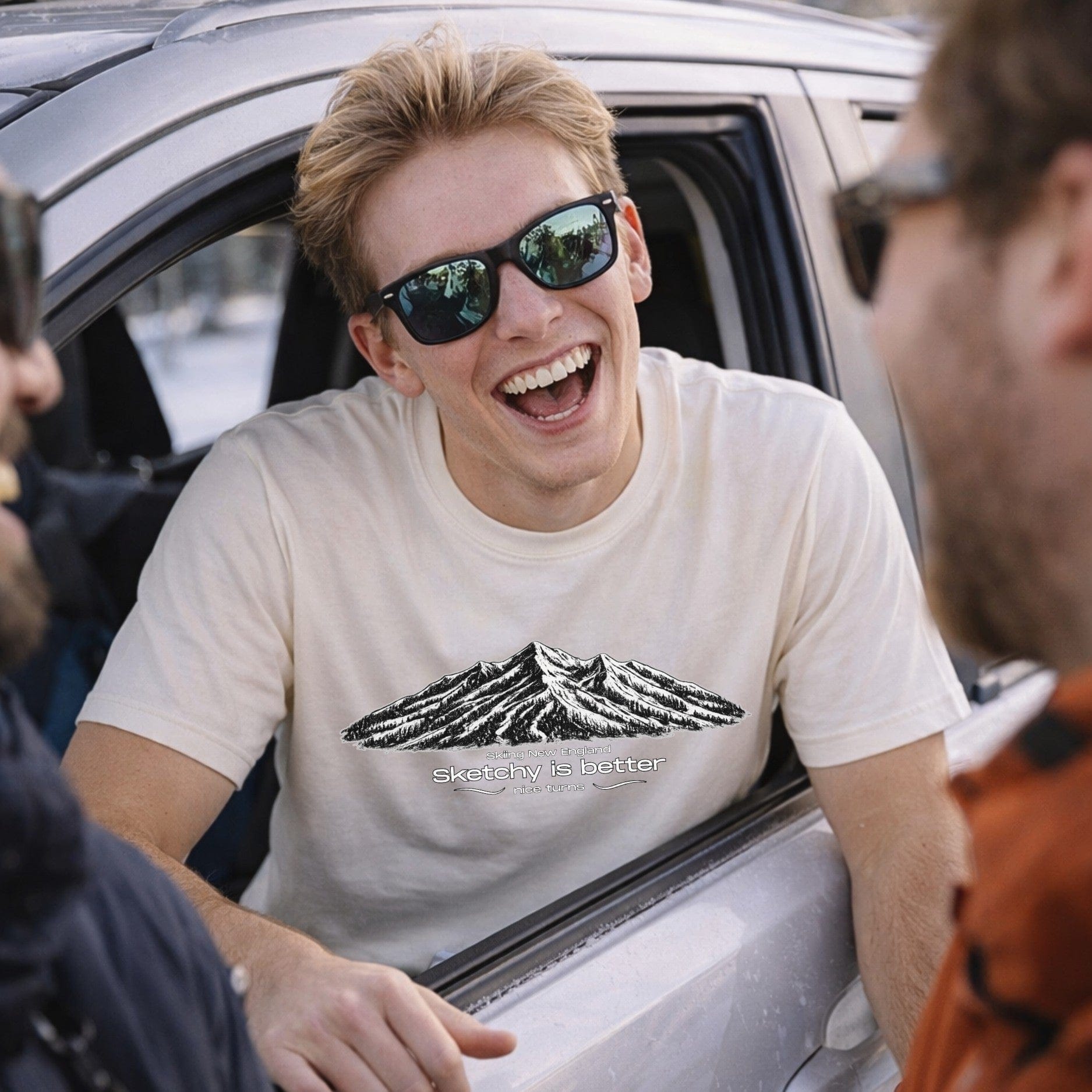 Male skier wearing cream mountain sketch t-shirt laughing near car après scene