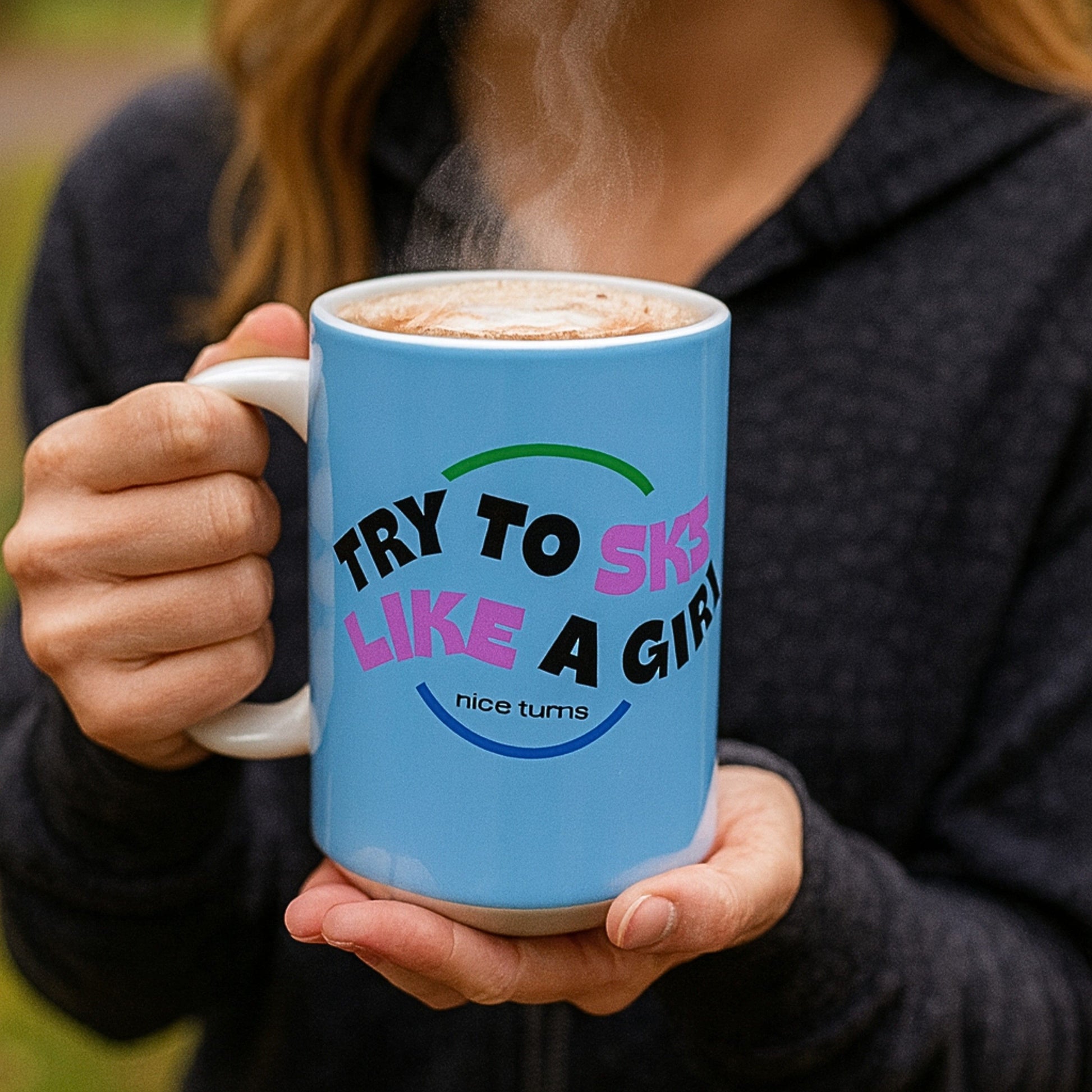 Freida Ridge 15 oz coffee mug, Cool Blue, held outdoors with steam rising; wraparound “Try to Ski Like a Girl” graphic clearly shown.