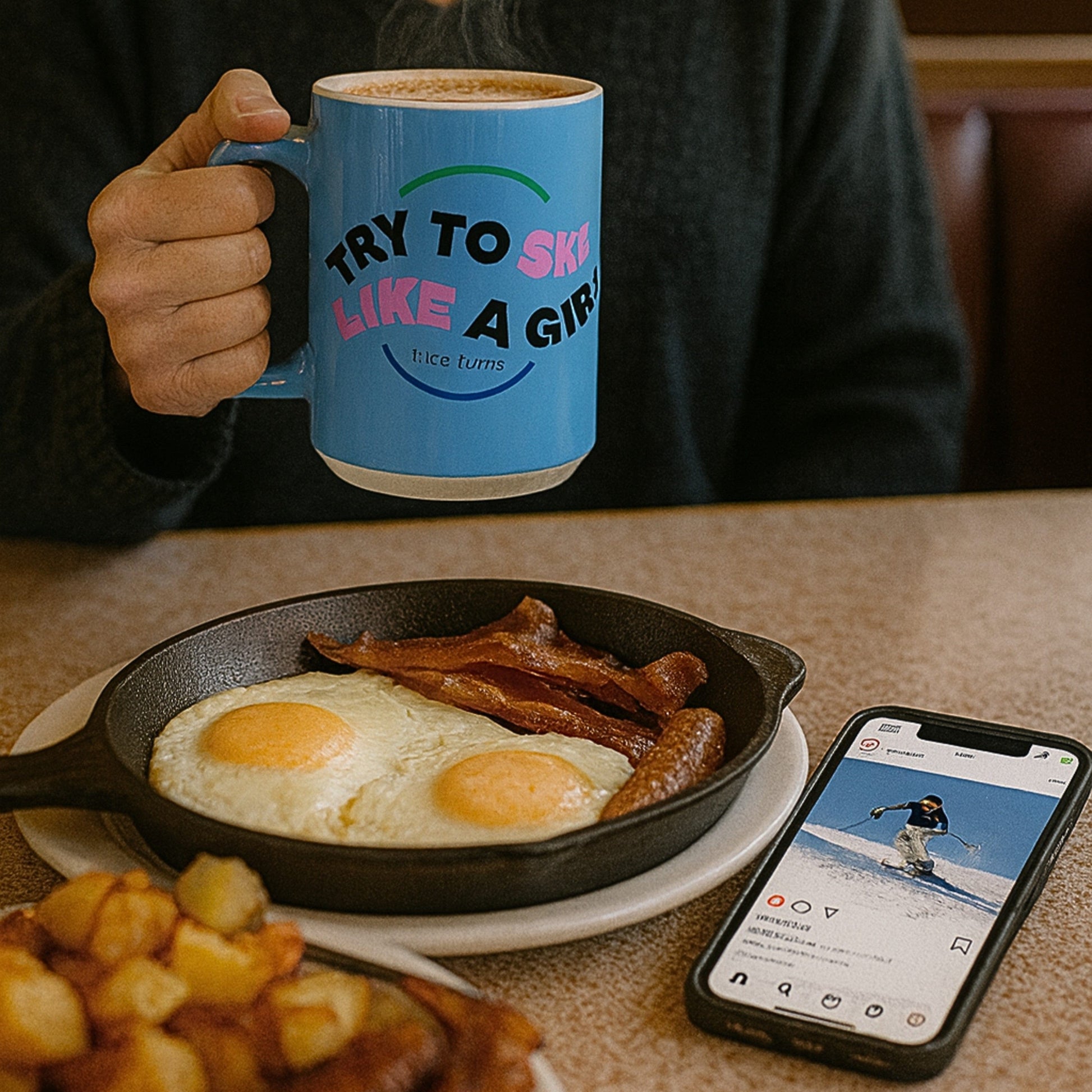 Freida Ridge 15 oz coffee mug, Cool Blue, held over diner breakfast with eggs and bacon; “Try to Ski Like a Girl” print clearly framed.