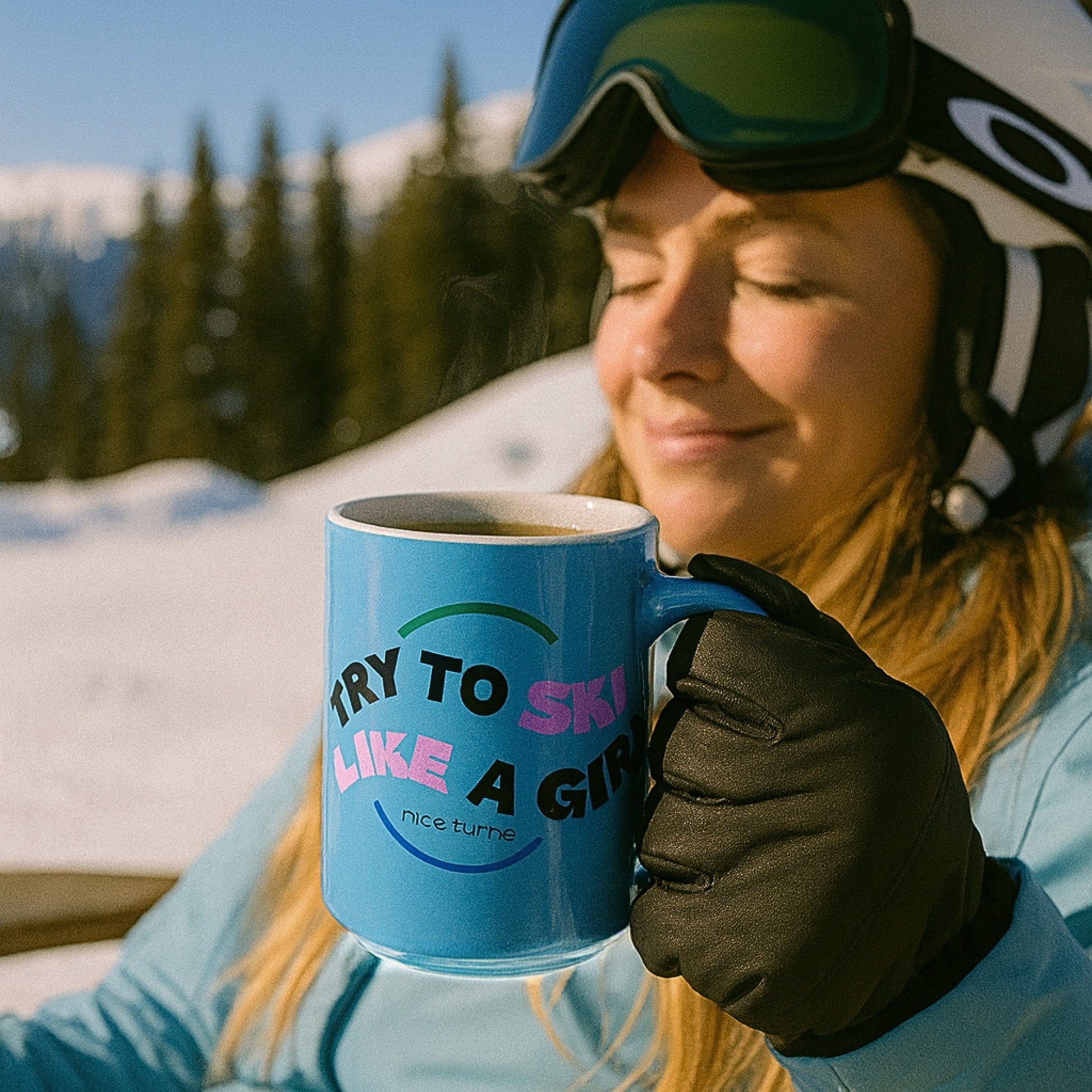Freida Ridge 15 oz coffee mug, Cool Blue, held by skier in gloves on snowy mountain deck; “Try to Ski Like a Girl” text visible with steam rising.