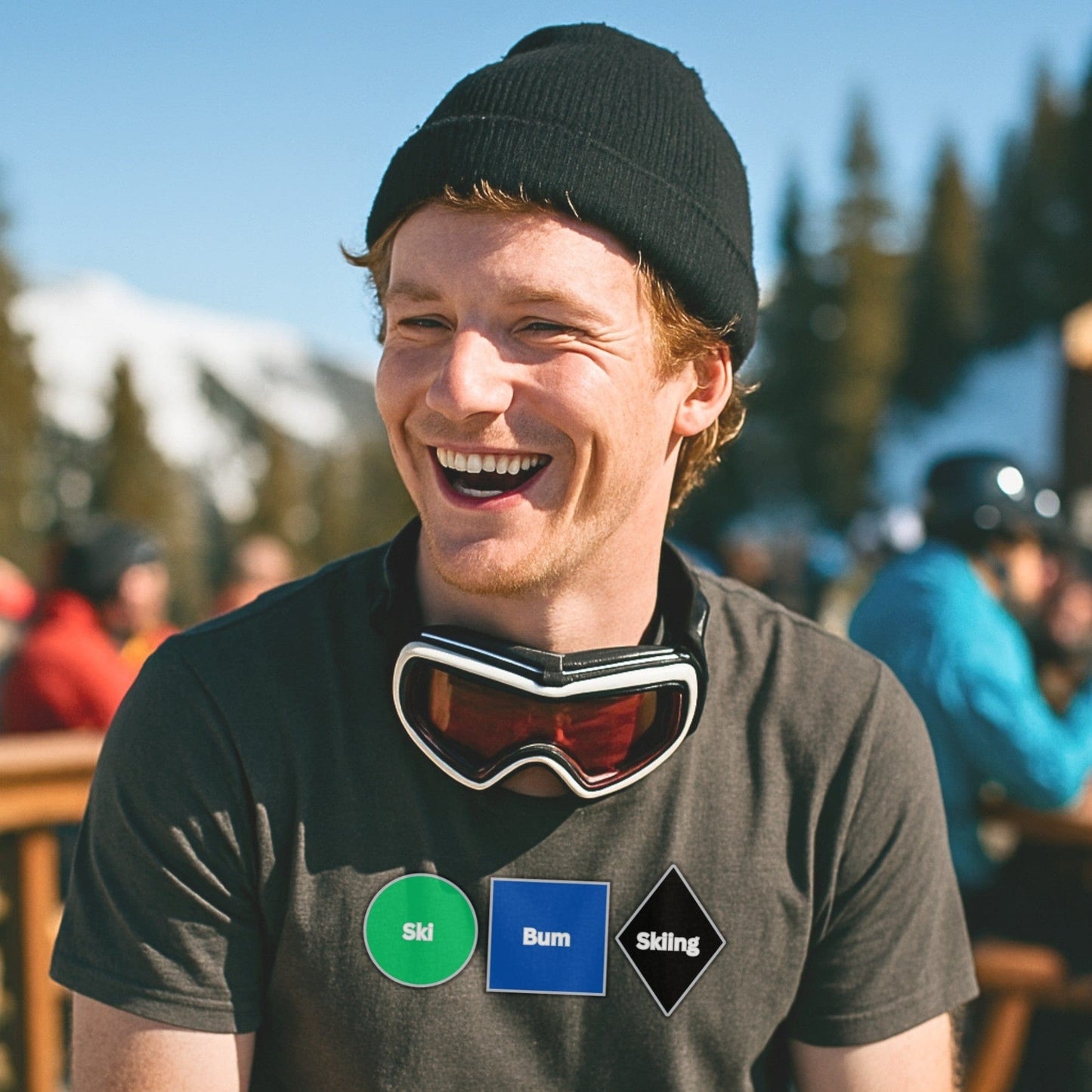 Smiling skier with goggles around neck wears No Lift Left Behind t-shirt in Heavy Metal gray on sunlit mountain deck, après scene in background.