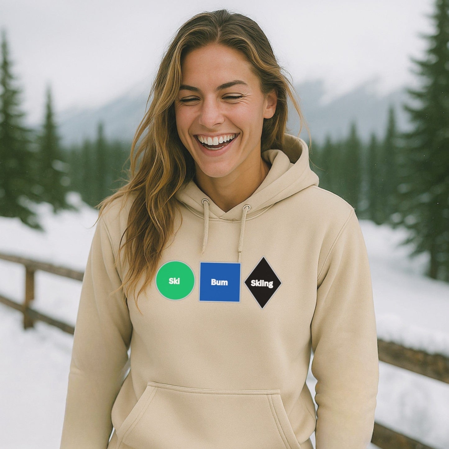 Sand-colored hoodie with Ski Bum Skiing design, worn by a smiling skier on a snowy mountain path lined with pine trees and a wooden fence.