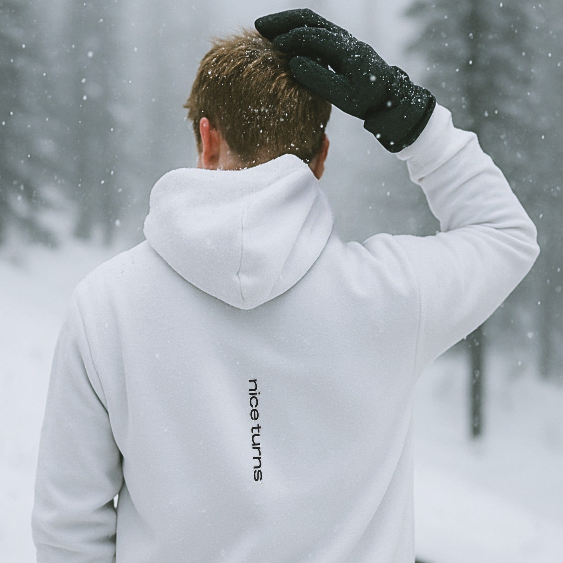 White hoodie with vertical nice turns logo on the back, shown in a snowy forest with falling flakes and soft winter light.