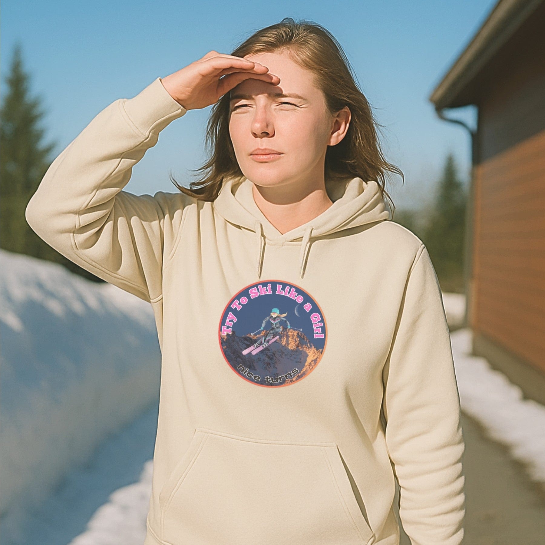 Sand-colored hoodie with Try to Ski Like a Girl graphic, worn by skier scanning the horizon near a sunny ski lodge with snowbanks.