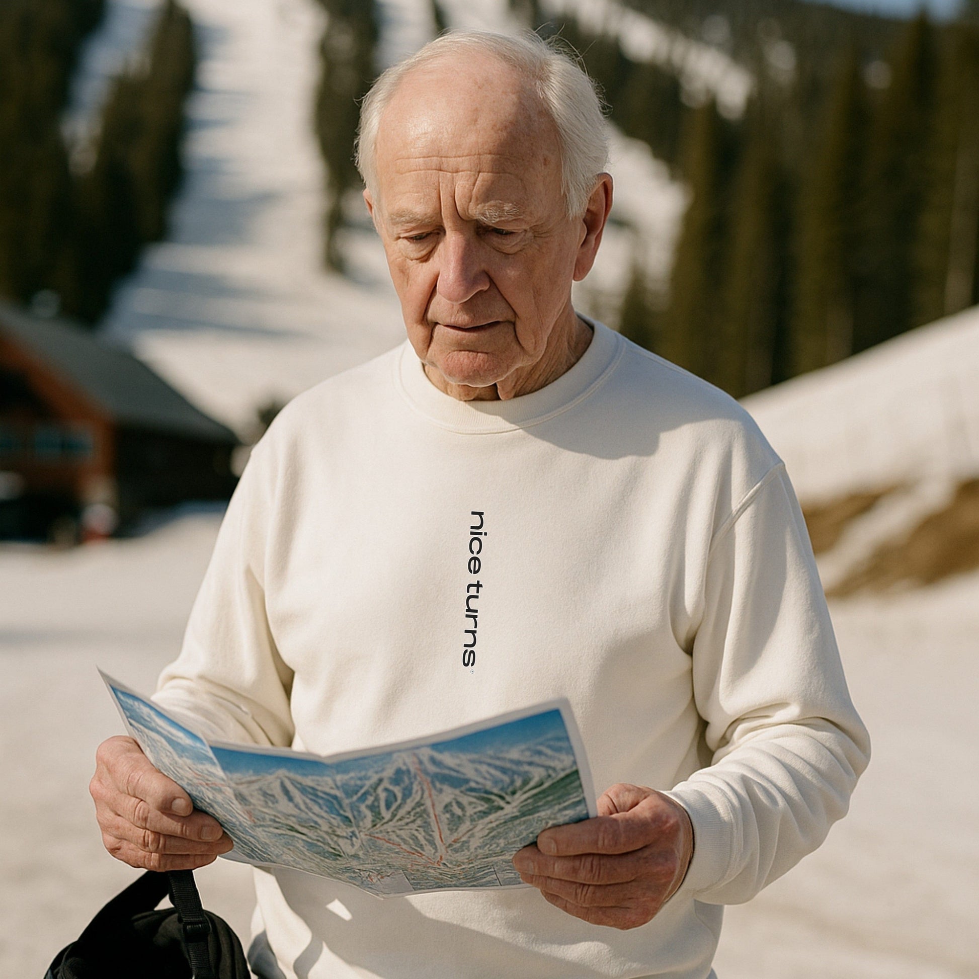 Bashing Bumps Sweatshirt, white, front lifestyle view on spring ski day; skier reading trail map with “nice turns” front mark visible.