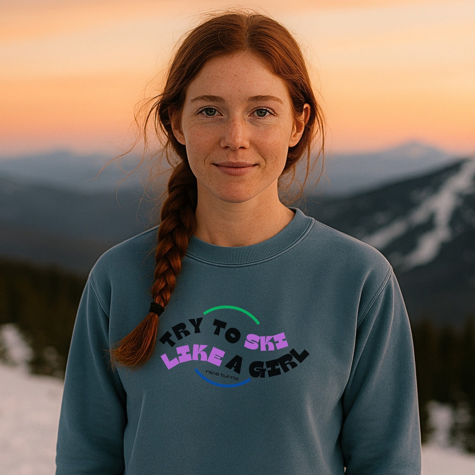 Woman with braided red hair wearing “Try to Ski Like a Girl” blue crewneck at sunset on snowy mountain ridge; retro-style lettering on front in Freida Ridge color.
