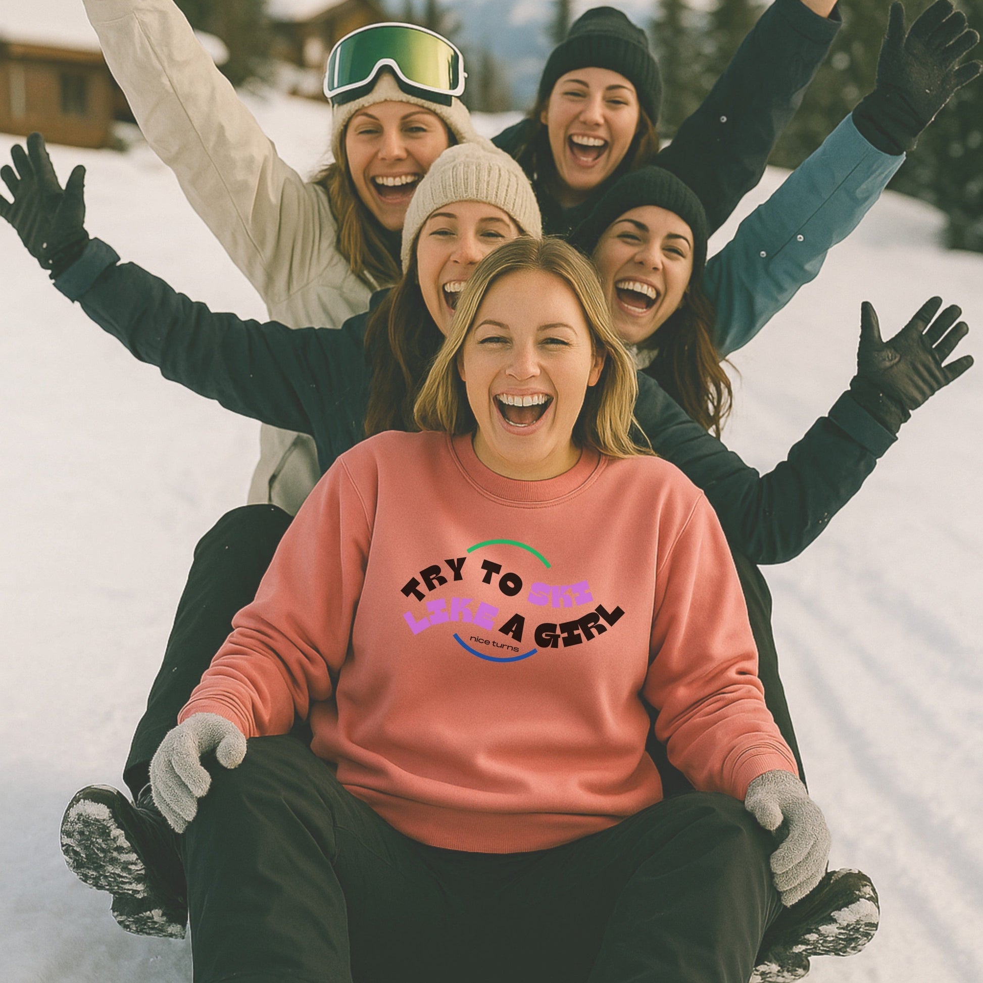 Five women laughing on a snow toboggan run, front rider in Watermelon “Try to Ski Like a Girl” crewneck; retro print pops against joyful winter backdrop.