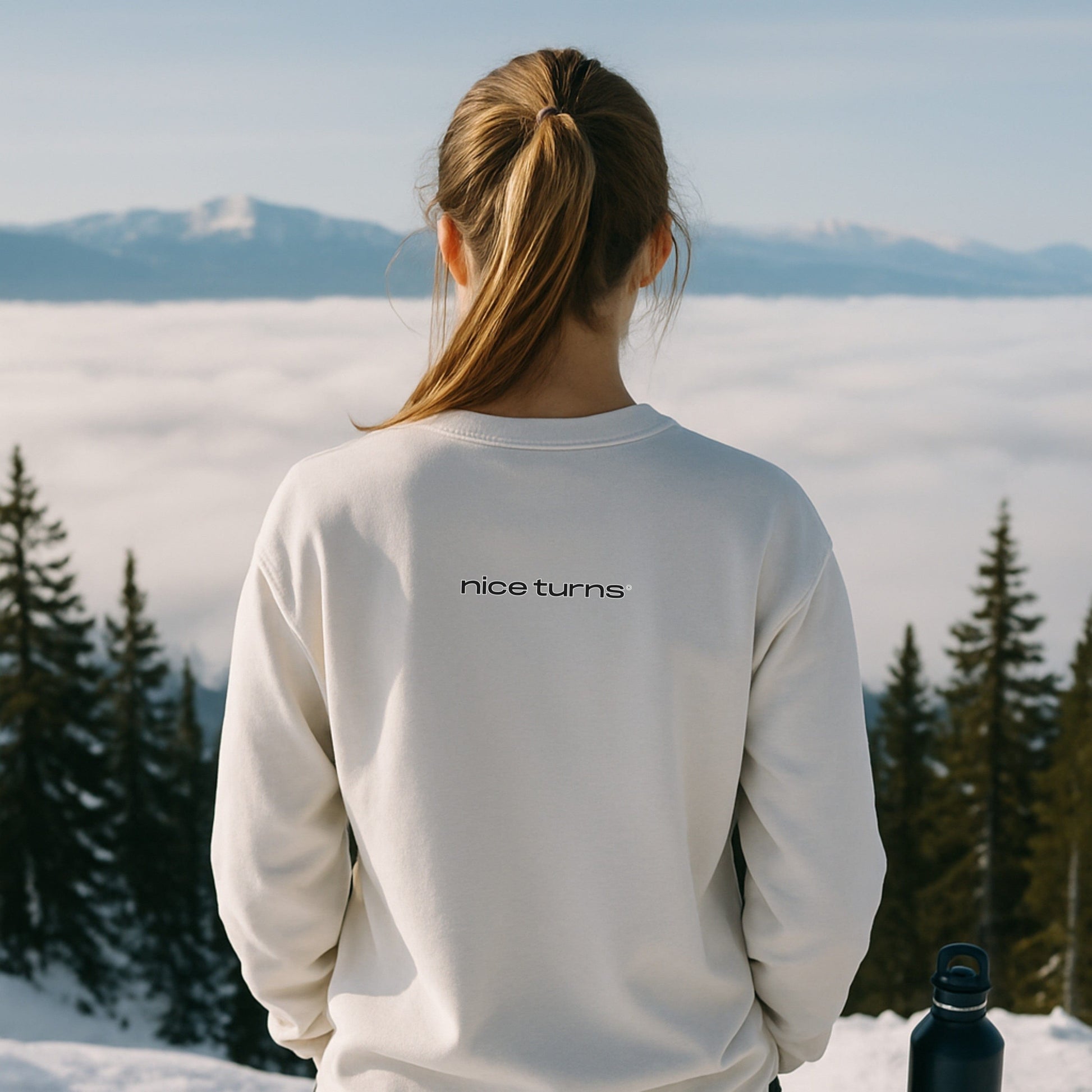 Back-view of skier in White crewneck with clean “nice turns” logo, gazing over a sea of clouds from the summit; ponytail over shoulder keeps the logo fully visible.