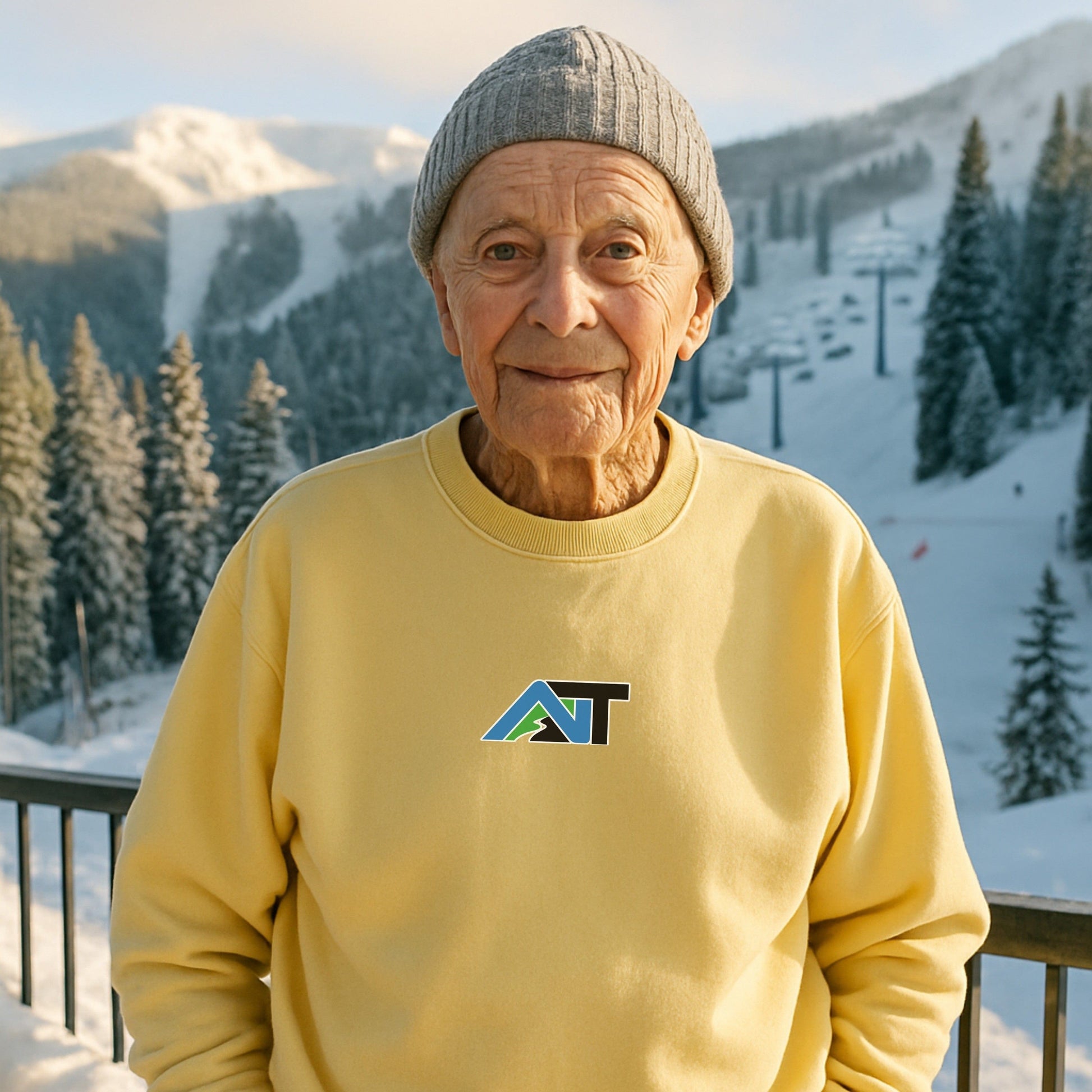 Elderly man in gray beanie and butter-yellow AT sweatshirt standing on snowy mountain overlook with ski slopes in background