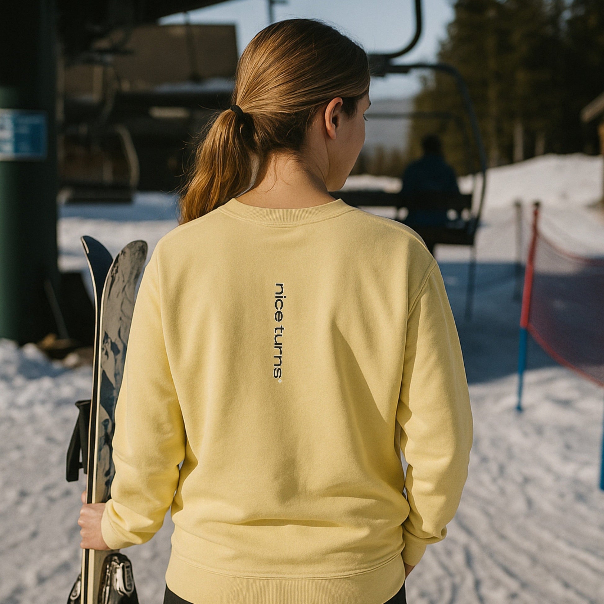 Back view of woman in butter-yellow sweatshirt with "nice turns" text, holding skis at the base of a sunny ski lift